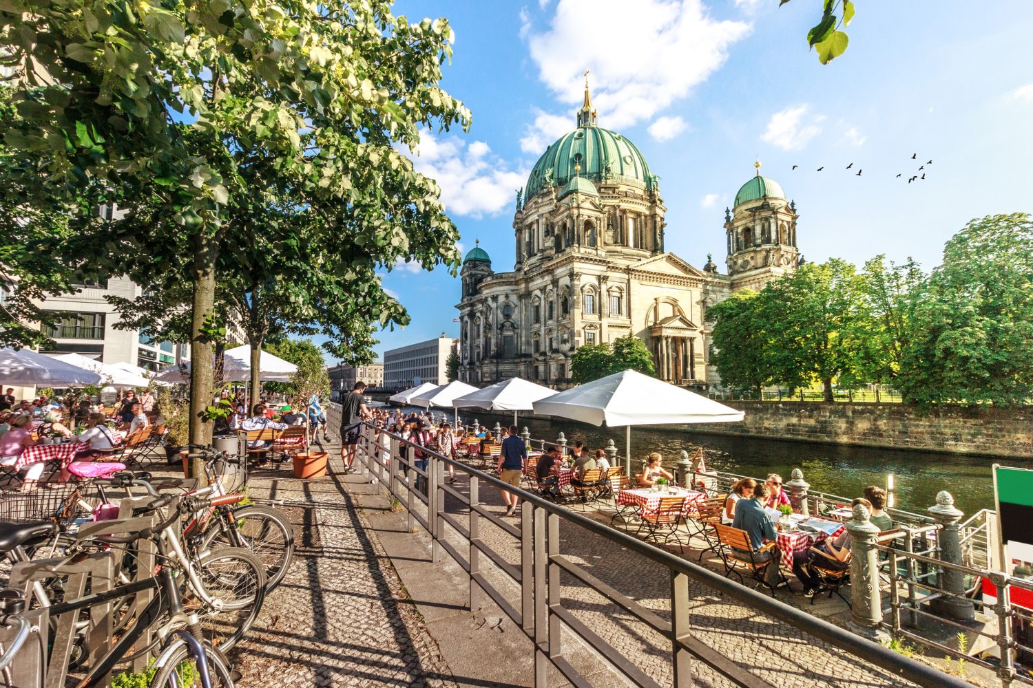 Outdoor cafe seating along a cobblestone riverside promenade with bicycles along the railing and a large domed cathedral in the background.