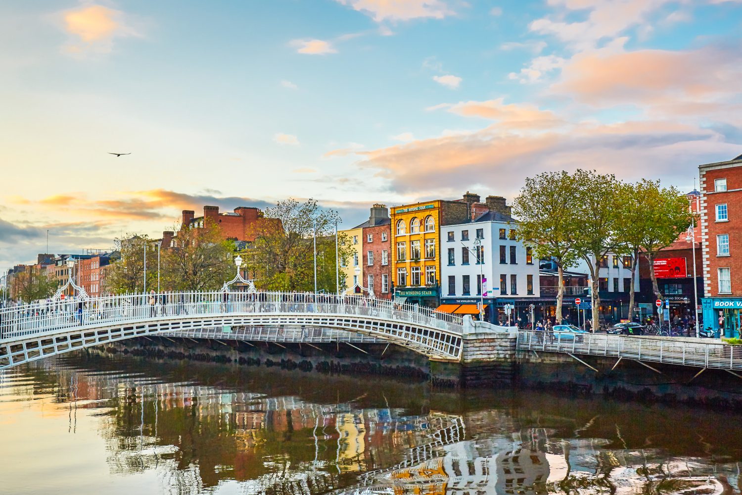 Colorful riverfront buildings line the bank with a white arched bridge spanning the water and trees reflected in the surface.