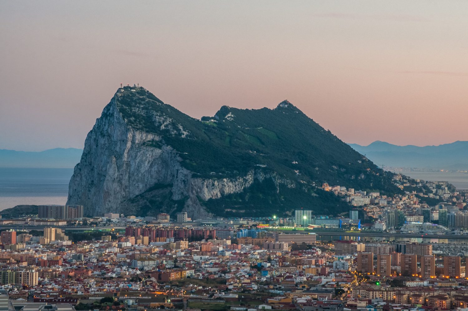Coastal city skyline at sunset with a large rocky mountain rising behind the buildings and the sea in the distance.