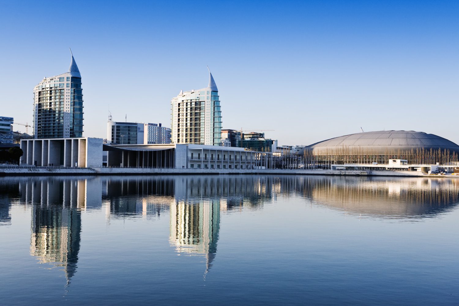 A modern city skyline along a calm body of water, featuring tall glass buildings and a large curved dome, with reflections on the surface.