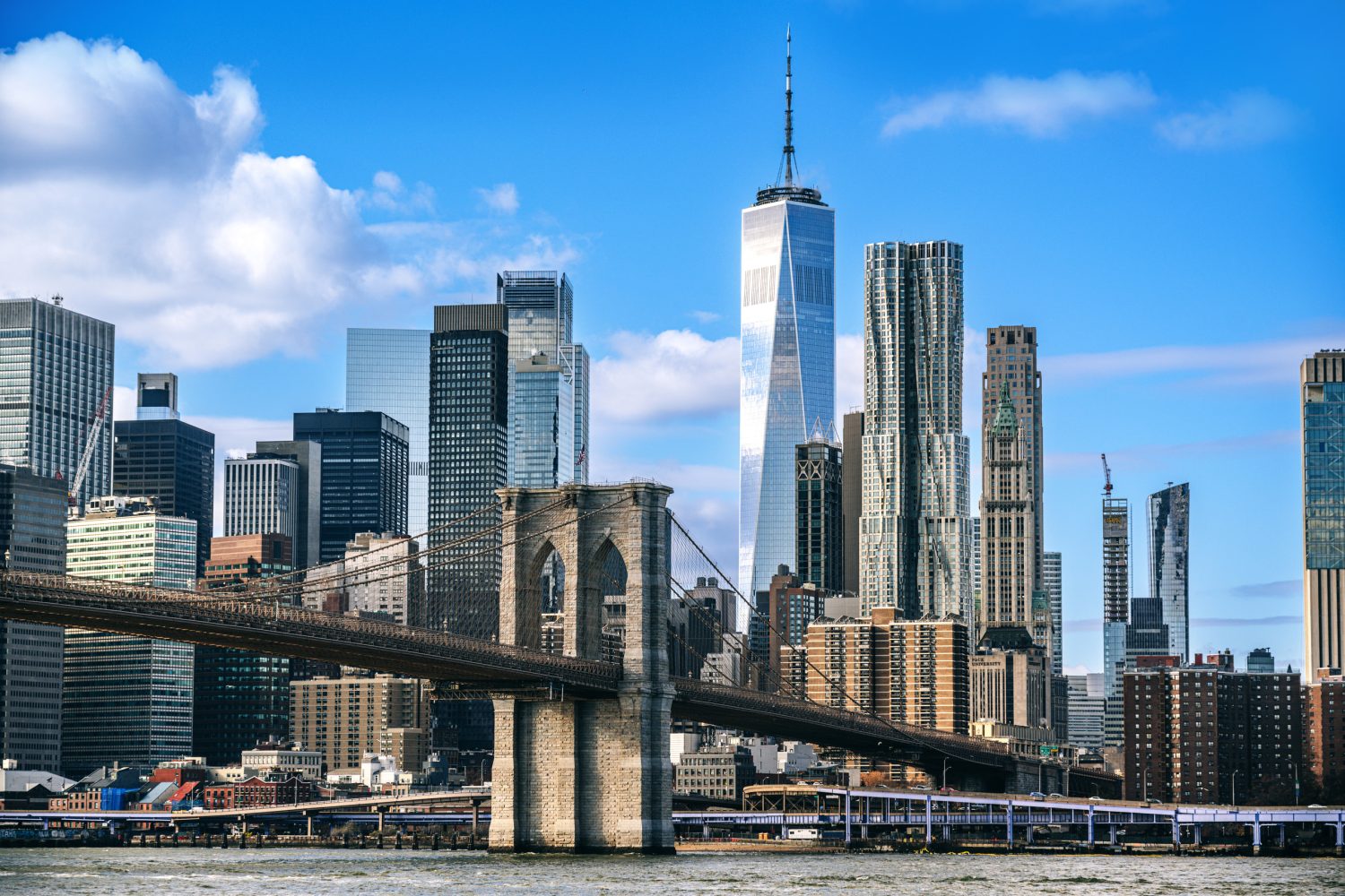 Brooklyn Bridge spans the East River with the Manhattan skyline, including One World Trade Center, rising behind it under a blue sky.