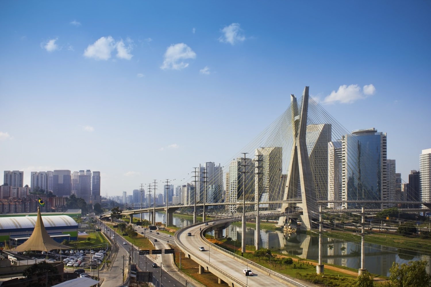 A modern city skyline with tall buildings, a curved bridge over a river, and a clear blue sky.