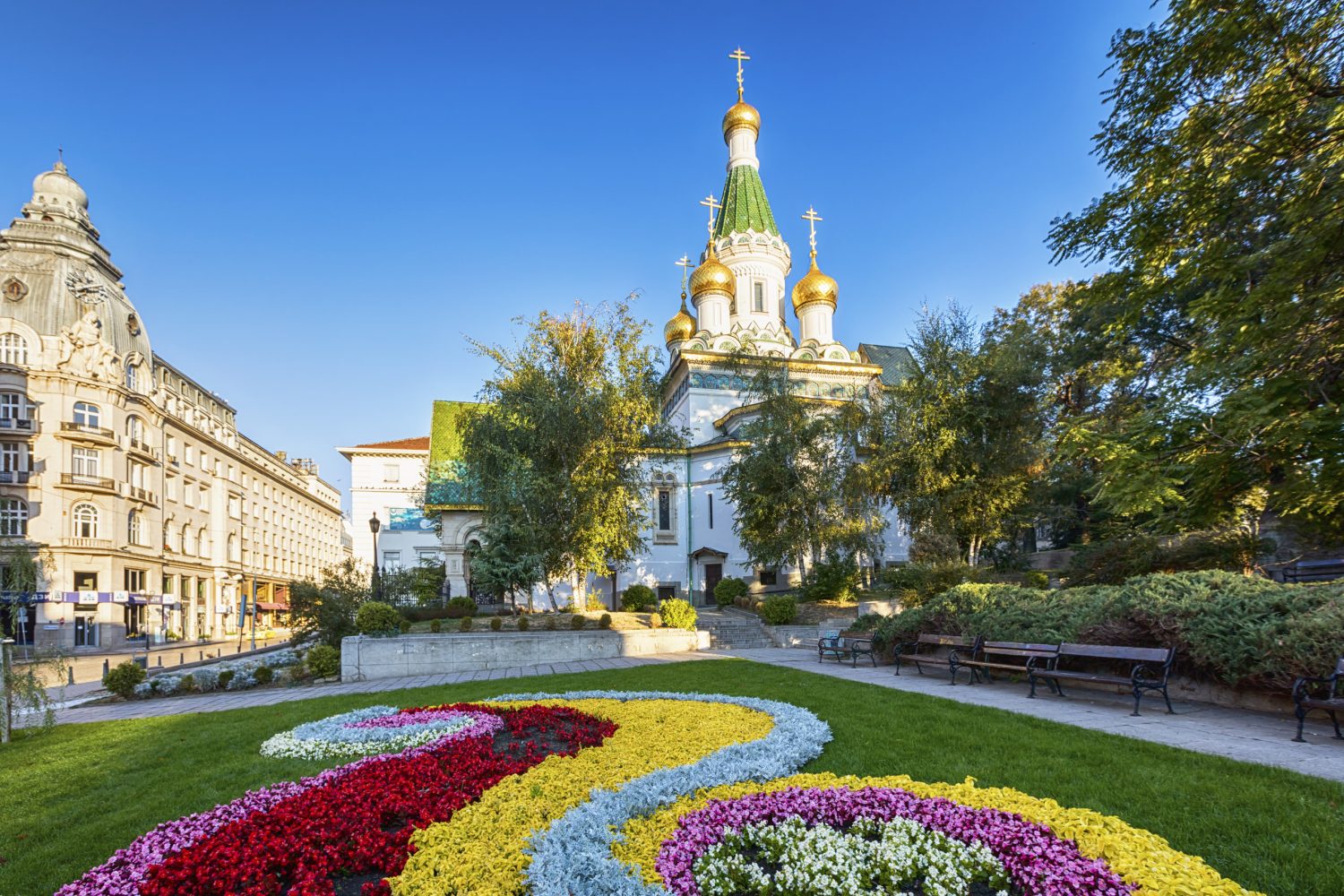 White Orthodox church with gold domes sits behind a vibrant circular flower garden in a sunny park.