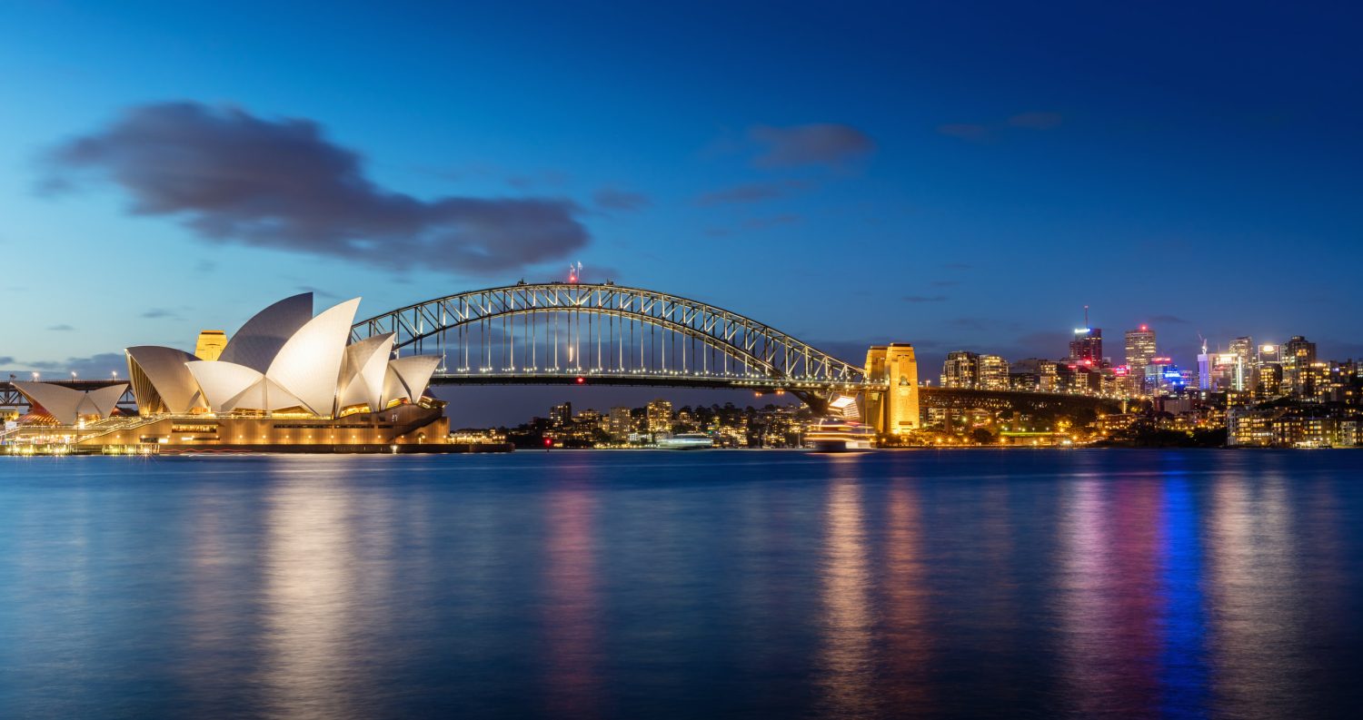 Nighttime view of the Sydney Opera House and Harbour Bridge illuminated along the waterfront with city lights reflecting on the water.