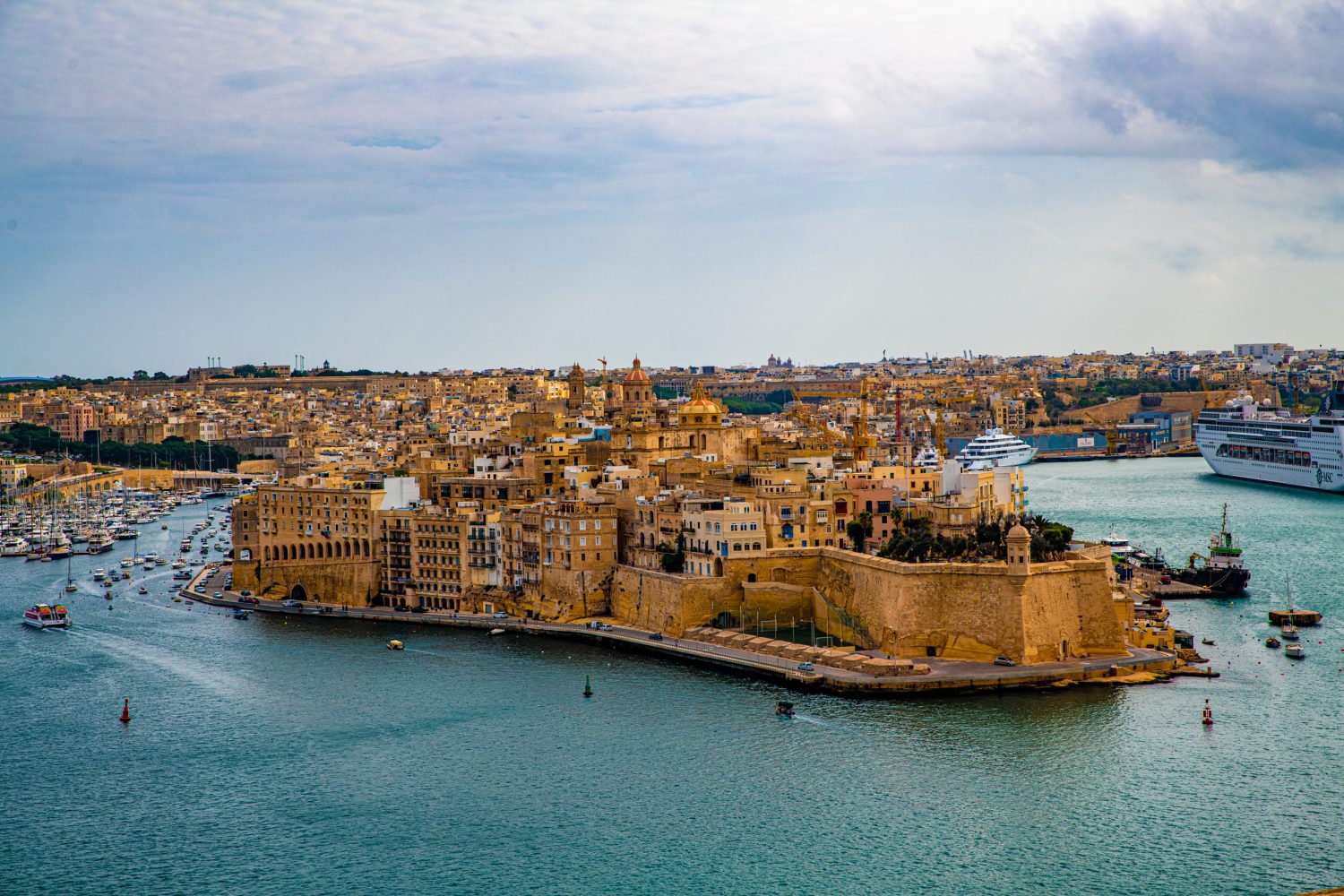 Historic fortress and tan buildings sit on a rocky promontory overlooking a calm blue harbor dotted with boats.