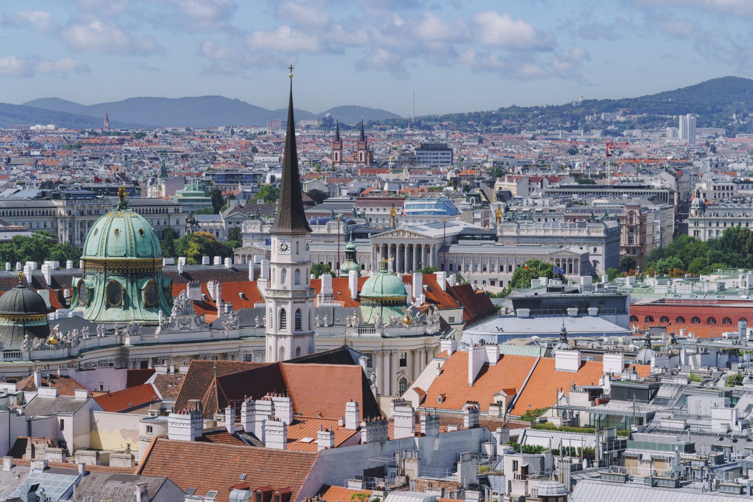 Panoramic view of a European city with red-tiled roofs, green-domed buildings, and a tall church spire against a distant skyline.