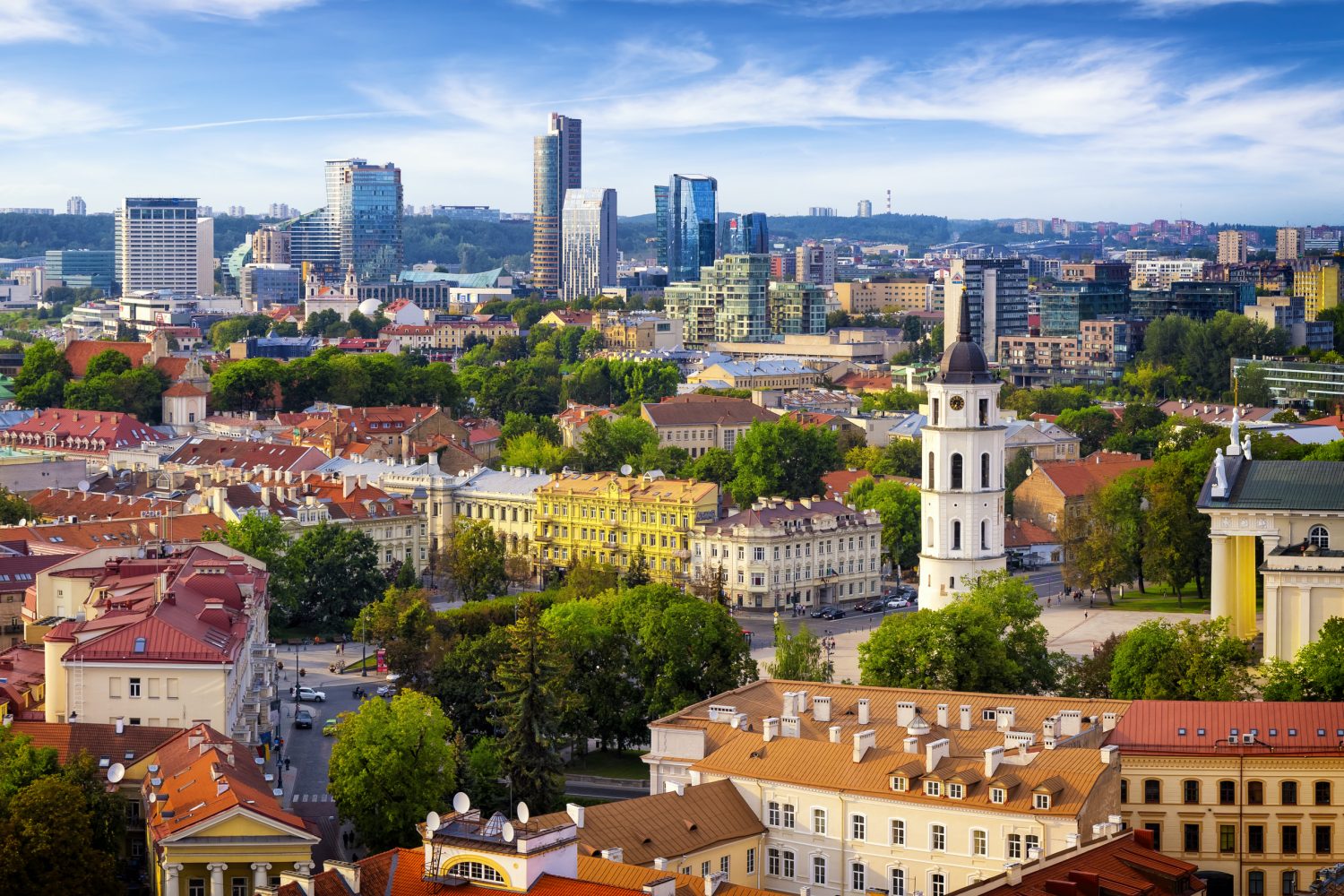 Panoramic cityscape of a European town with colorful rooftops, a prominent white church tower, green trees, and a blue sky.