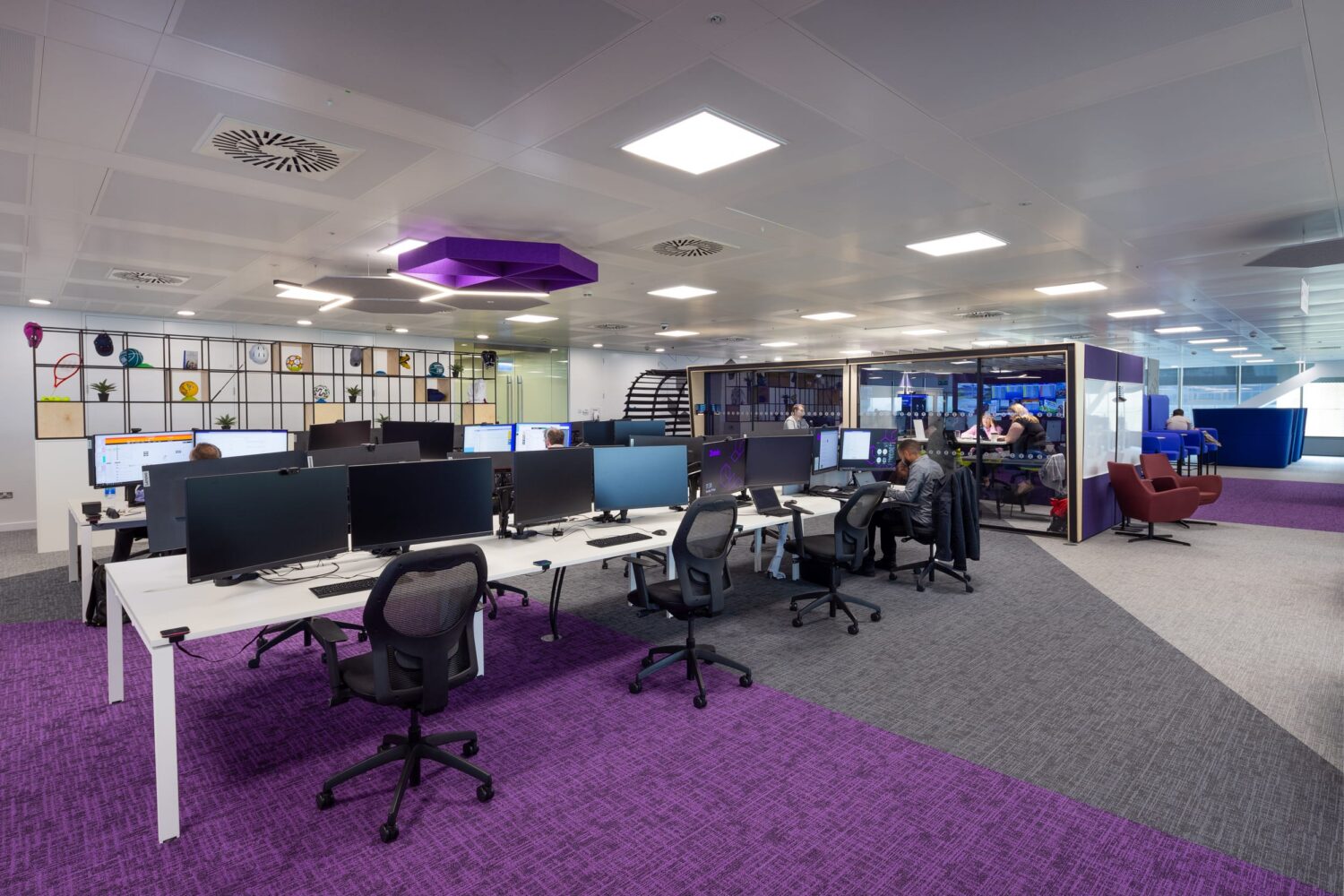 Modern open-plan office with rows of desks, computer monitors, and a purple-carpeted floor, plus a lounge area in the background.