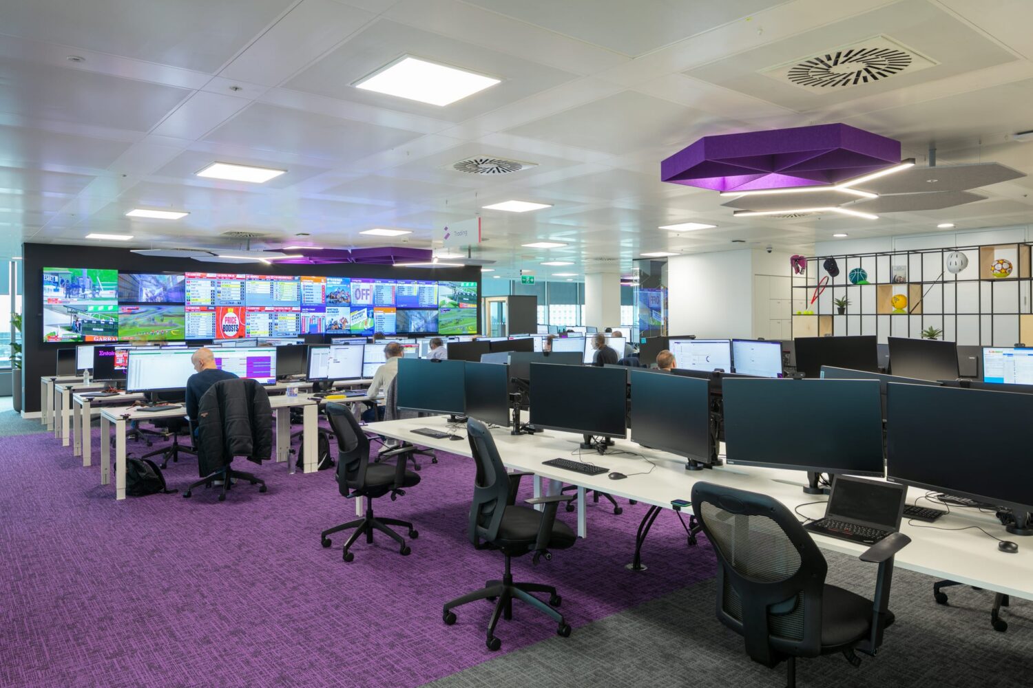 A modern control room with rows of desks and computer workstations facing a large wall of video screens, set on a purple carpet.