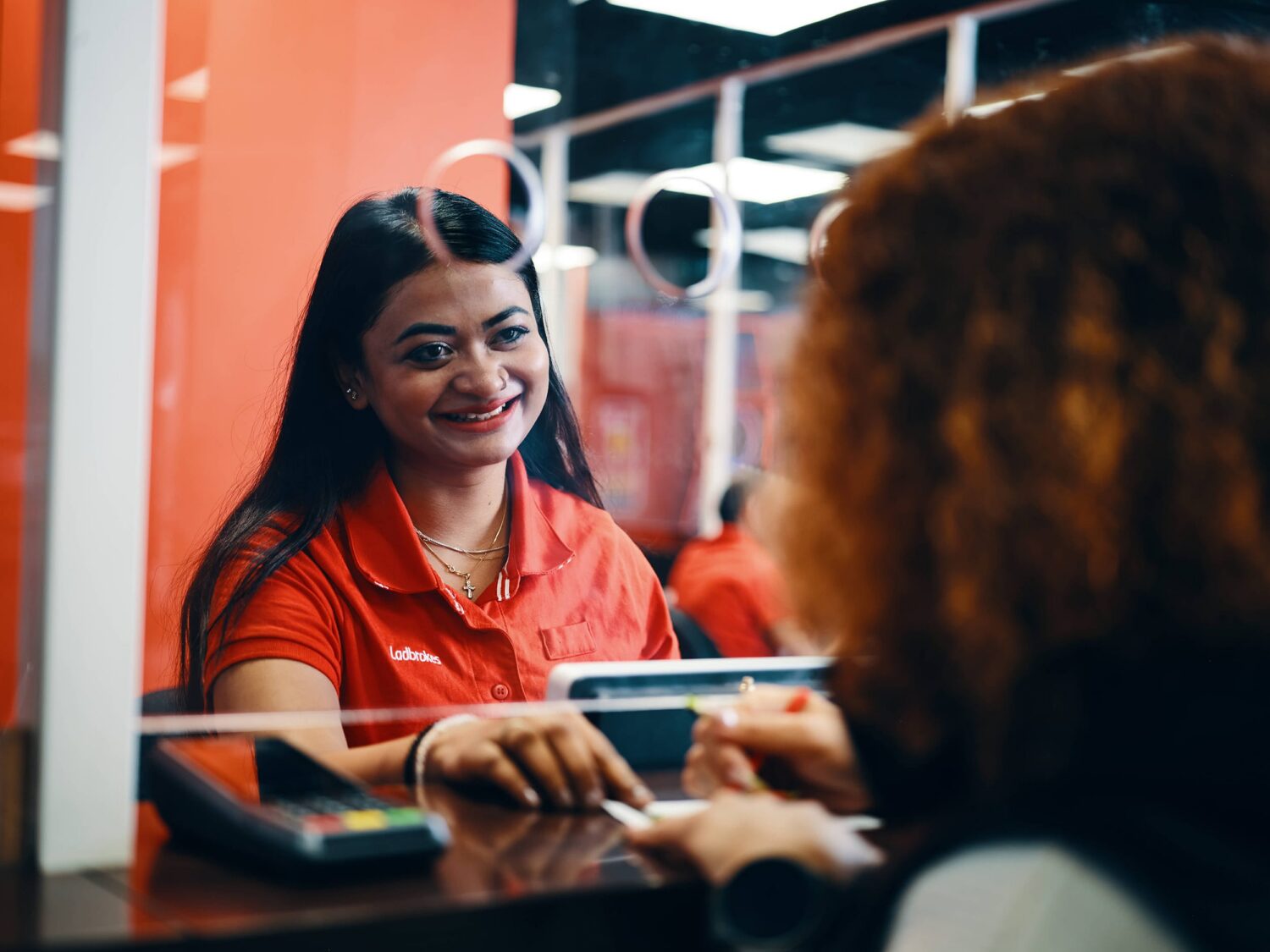 Smiling woman in a red polo greets a customer at a front desk with a card payment terminal.