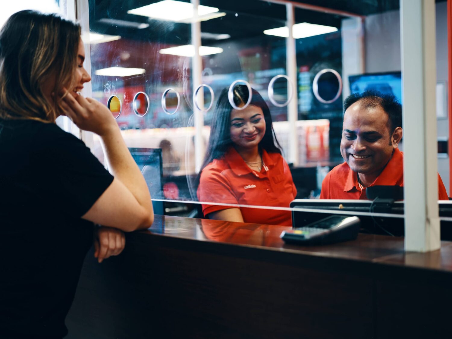A customer at a service counter talks to two smiling staff behind a glass partition.