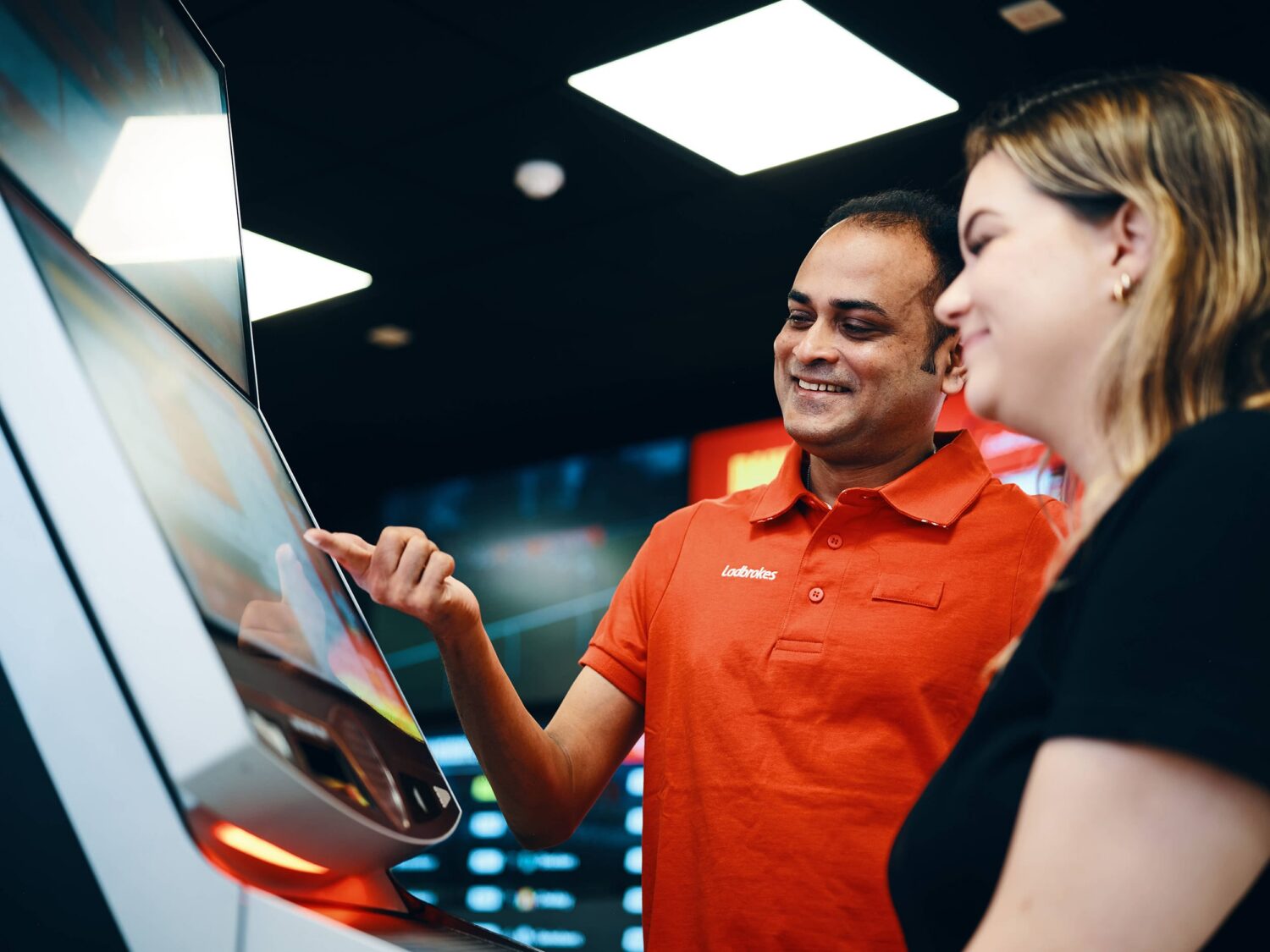 A man in an orange polo and a woman smile as they use a touchscreen kiosk.