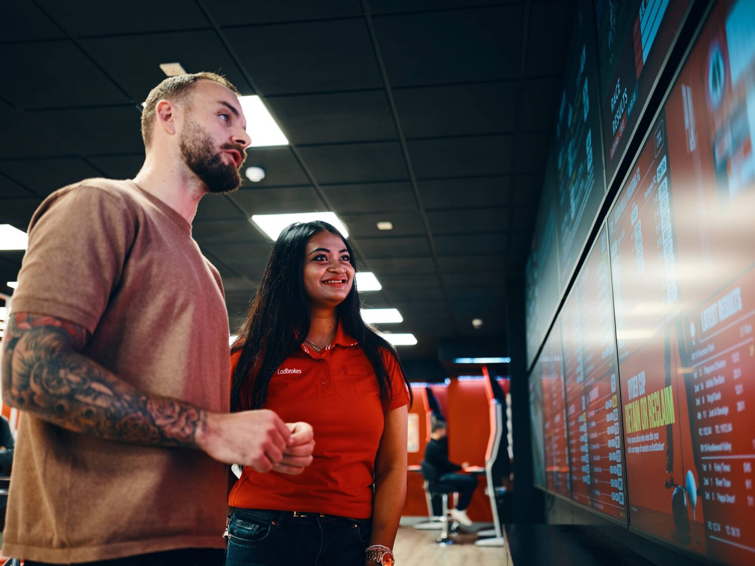 Man with a beard and a woman in a red shirt stand side by side, looking at a large illuminated touchscreen display on the wall in a modern indoor space.