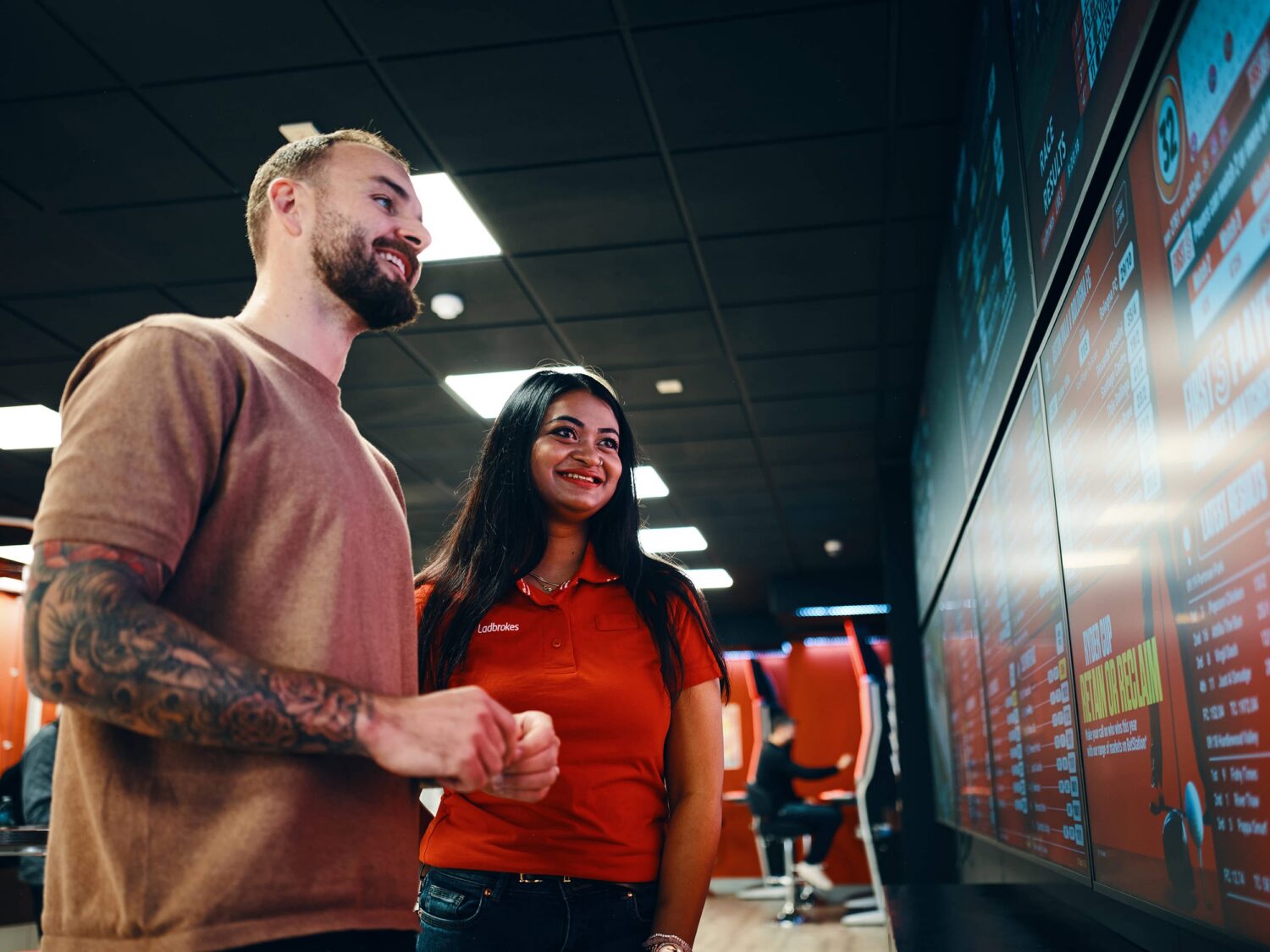 A bearded man with a tattoo sleeve and a woman in a red top smile as they look at a large wall-mounted touchscreen display in a modern indoor space.