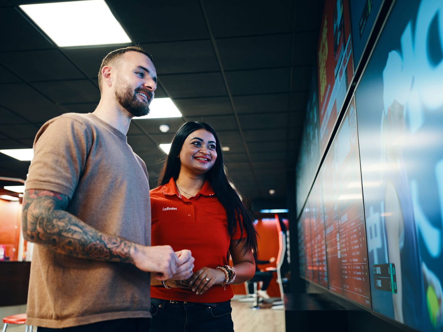 A man with a tattooed arm and a woman in a red shirt smile as they look at a large touchscreen display in a store.
