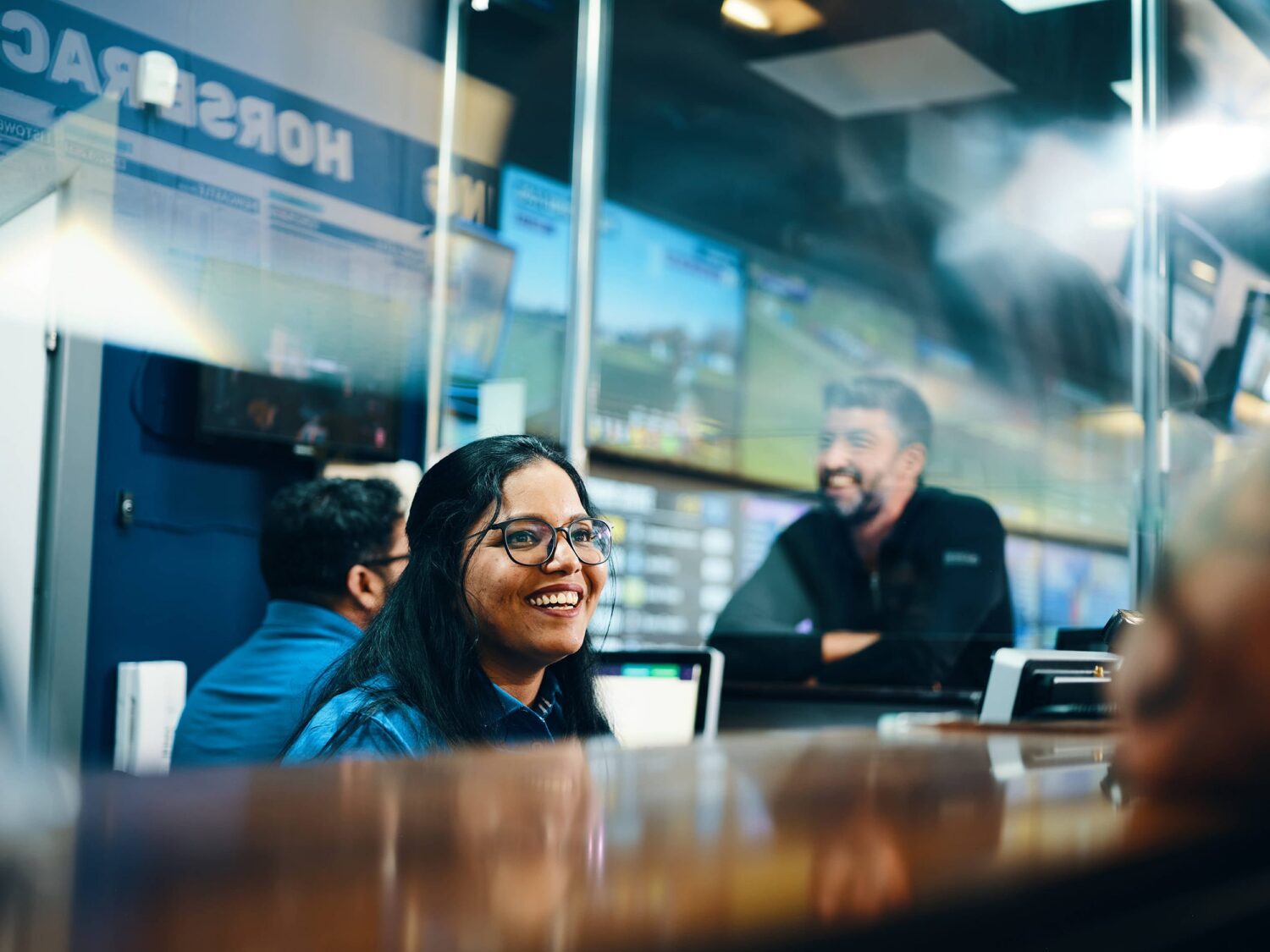 A smiling woman wearing glasses sits at a desk inside a glass-walled office, with coworkers and screens visible in the background.