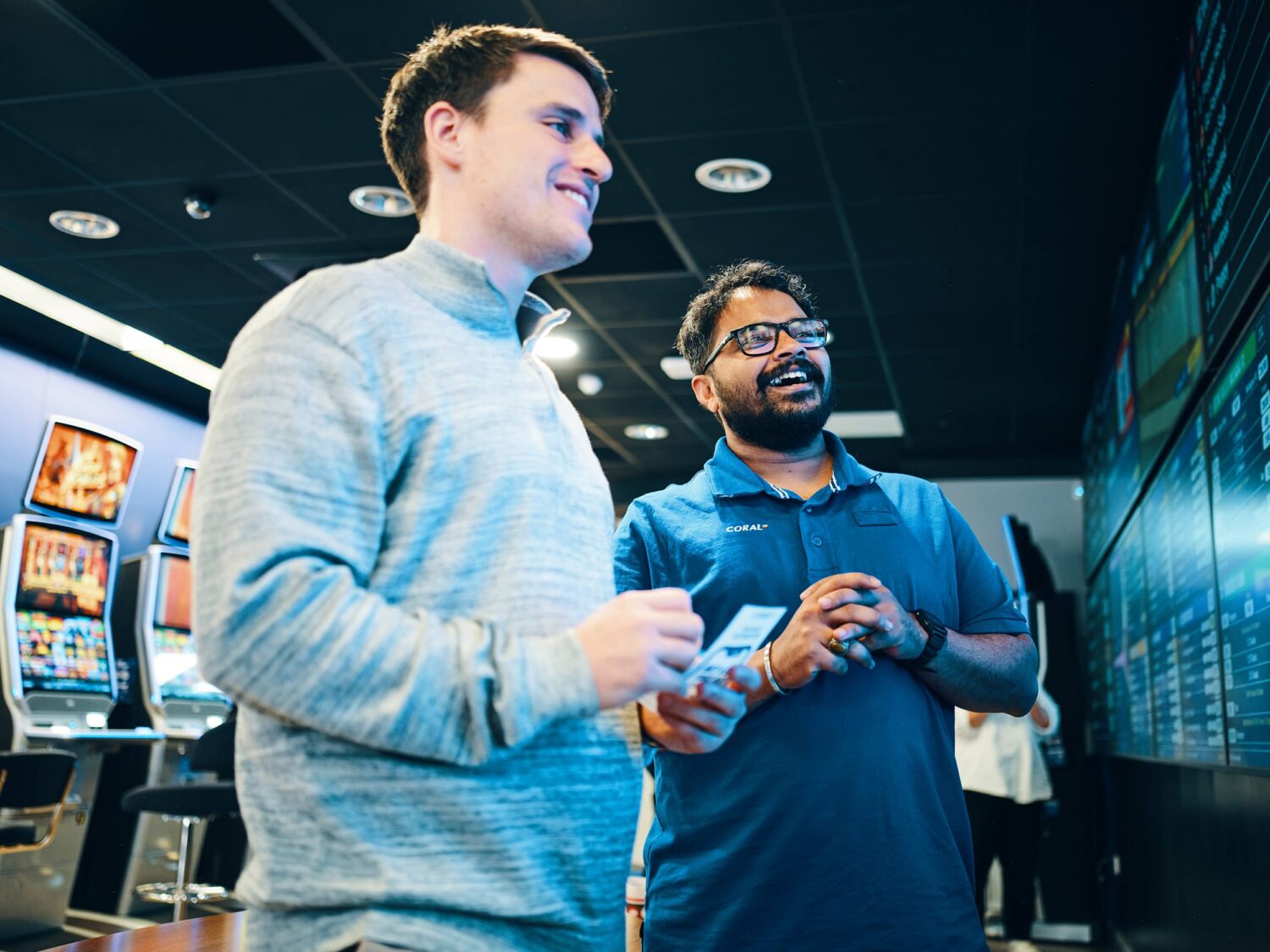 Two men in casual clothing stand in a gaming arcade near a wall of digital screens, smiling.