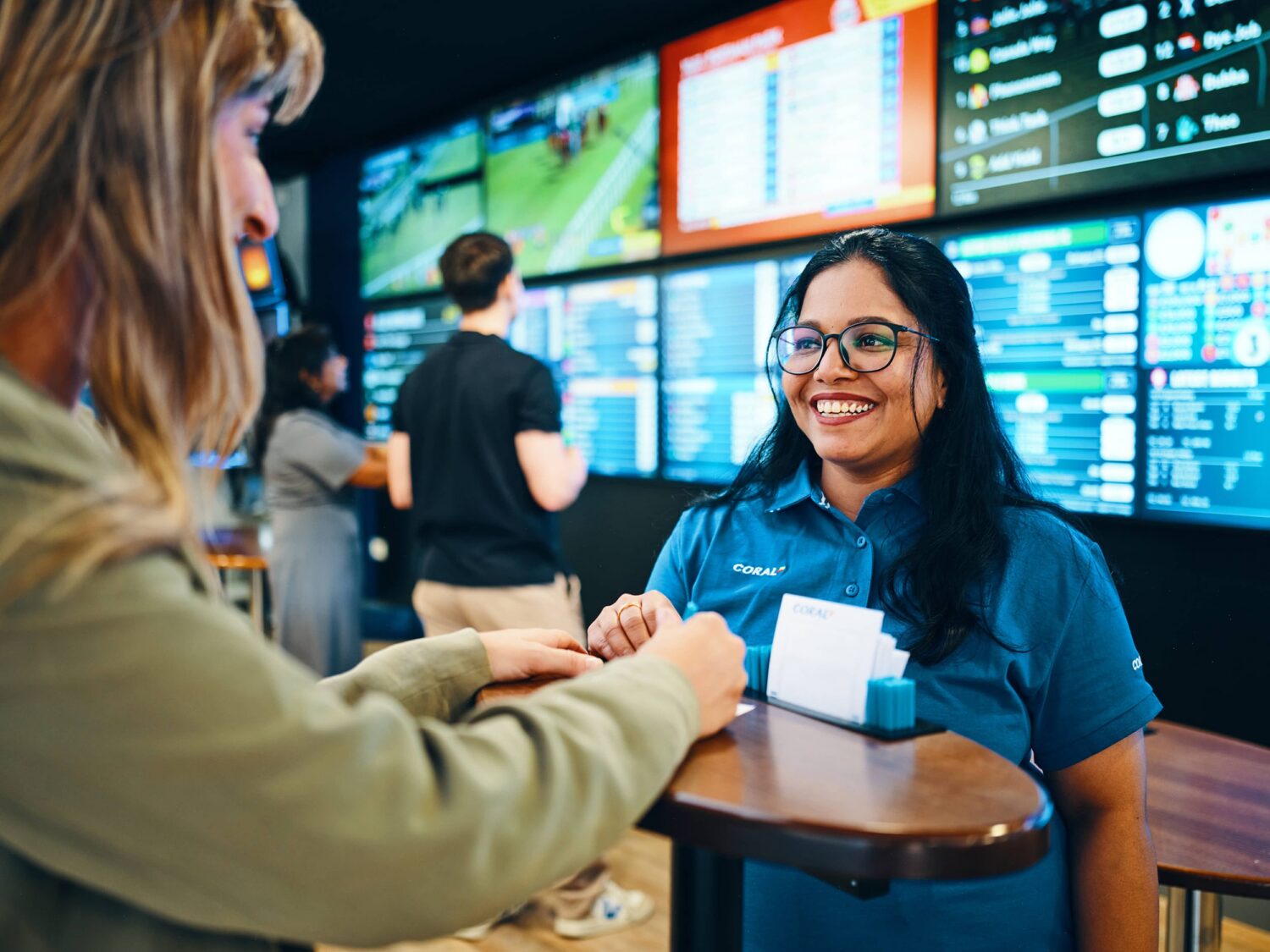 Smiling service agent in a blue uniform assists a customer at a counter with large screens displaying data in the background.