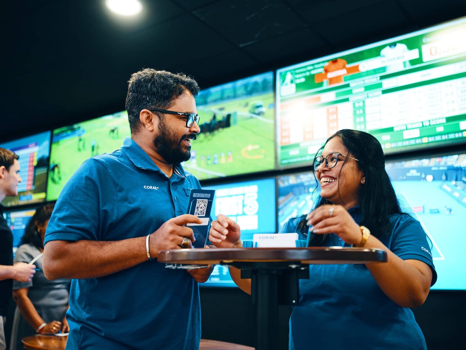 Two colleagues in blue polo shirts smile while sharing coffee on a tray in a high-tech control room with multiple screens.