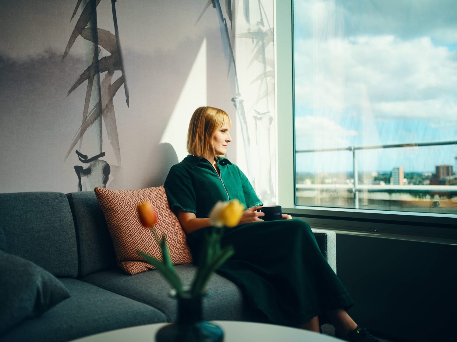 A woman sits on a gray sofa by a large window with a city view, holding a cup, while a vase of orange flowers rests on a nearby table.