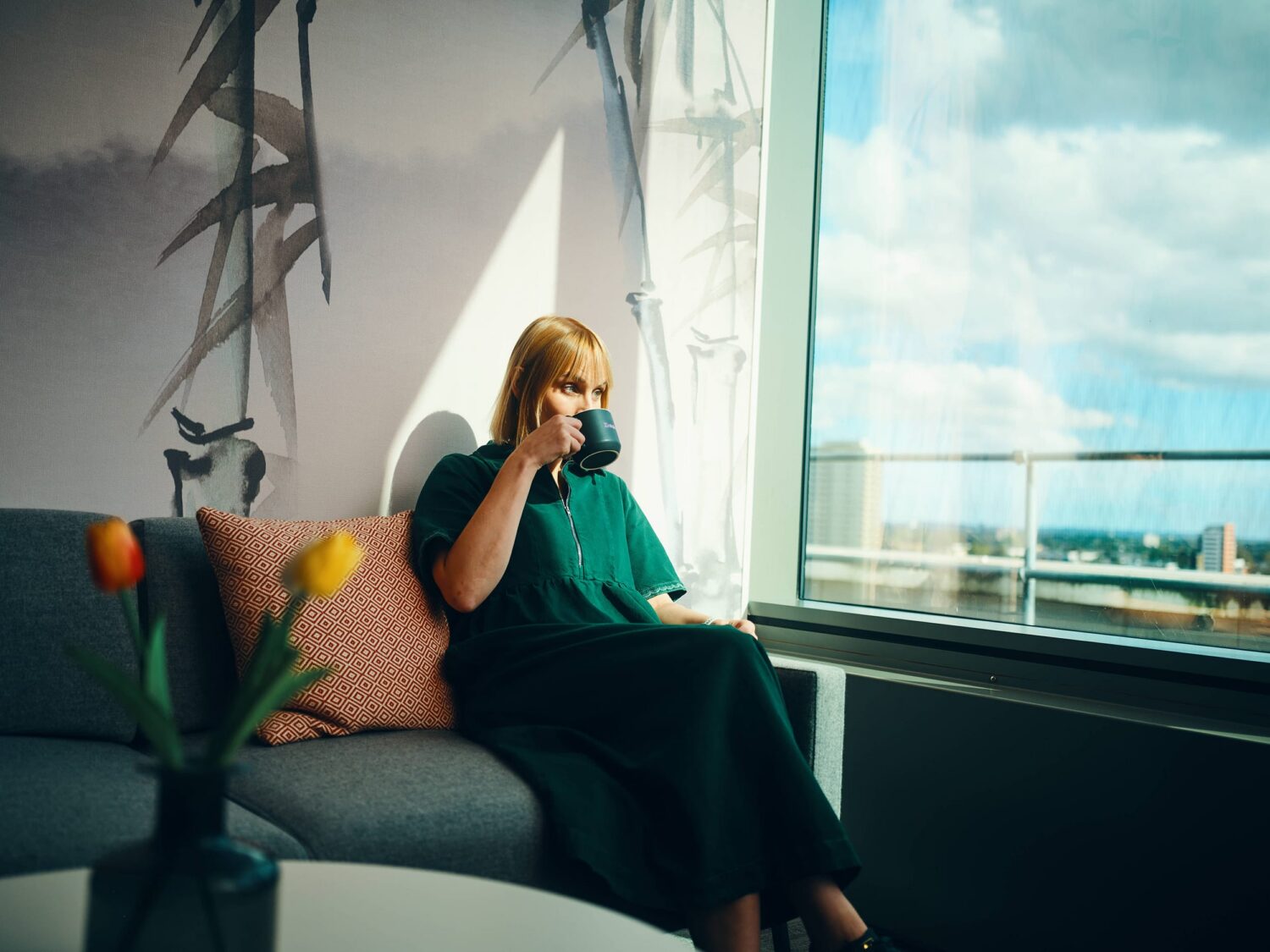 A woman in a green dress sits on a gray sofa by a large window, sipping from a mug, with a vase of tulips on a nearby table.