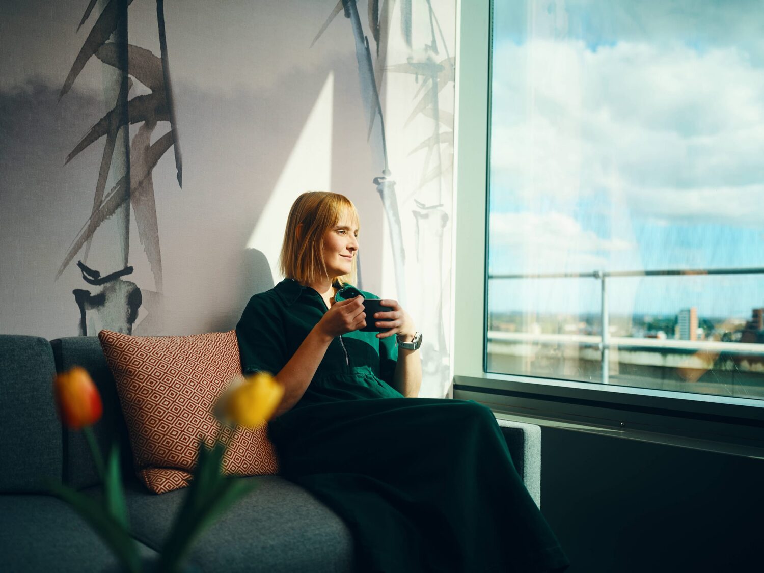 A woman sits on a sofa by a large window, holding a mug and looking out at a water view.