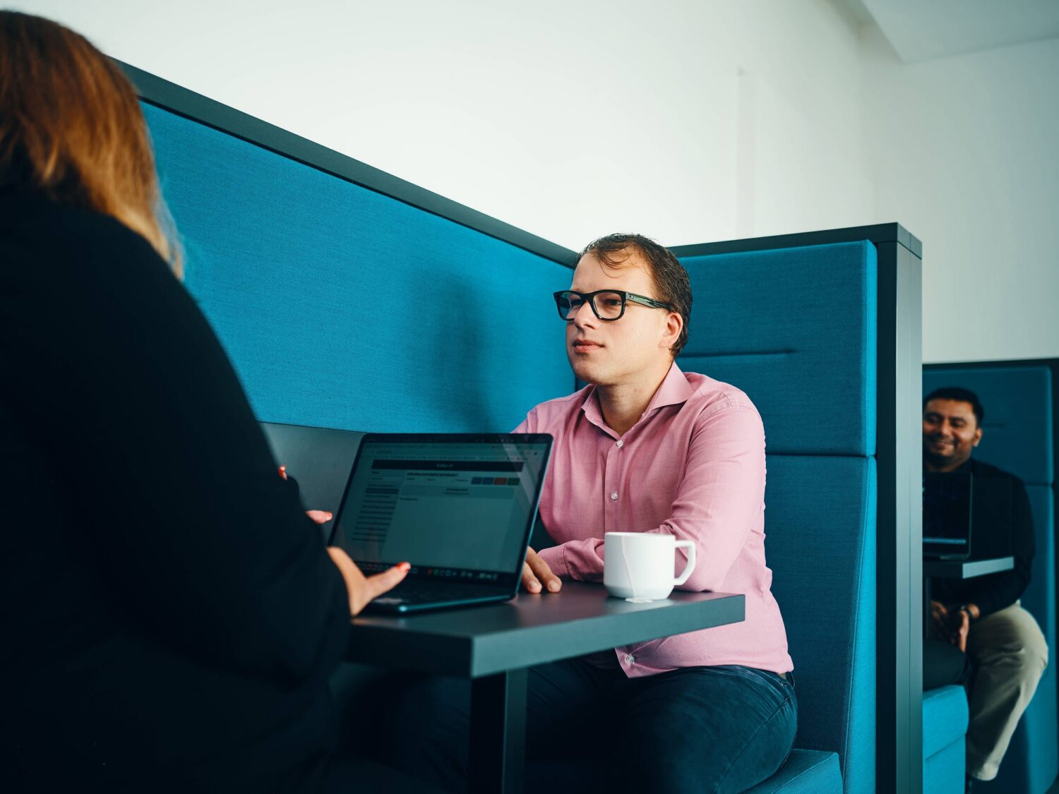 Man in a pink shirt sits at a table in a blue booth with a laptop and mug, talking to another person.