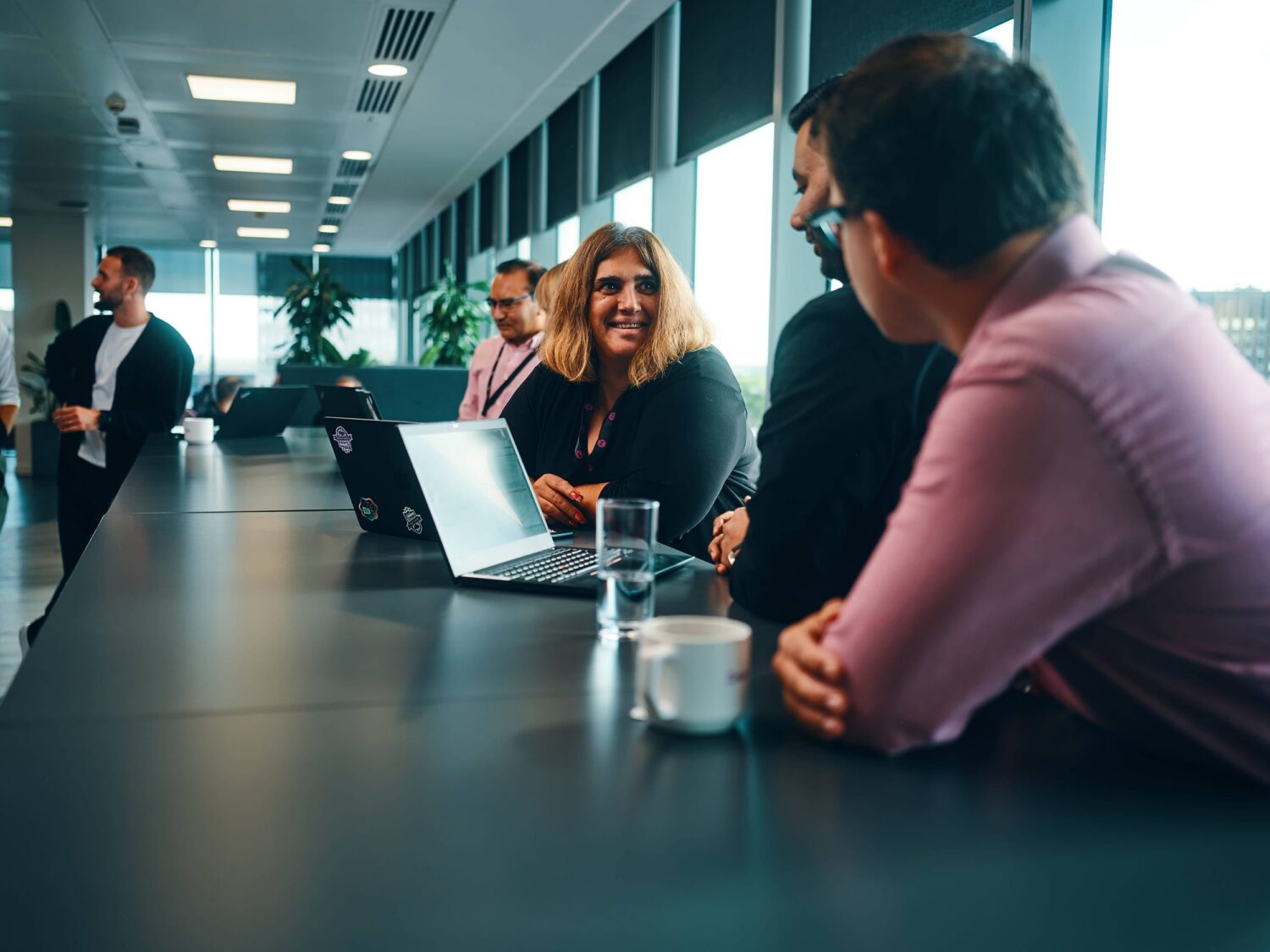 Colleagues sit around a long conference table in a bright office, smiling at a laptop.