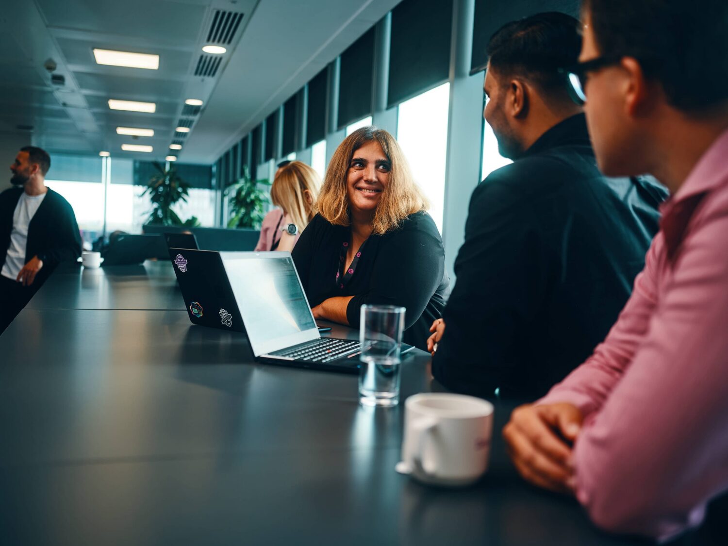 A smiling woman sits at the center of a long conference table in a bright office, surrounded by colleagues with laptops.