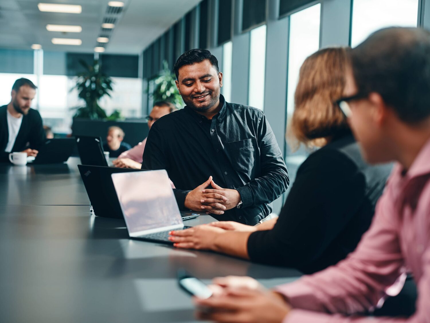 A group of colleagues collaborates around a conference table with laptops in a bright, modern office.