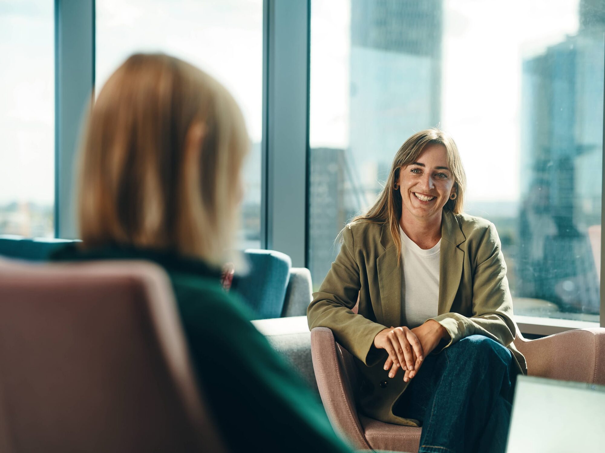 Two women sit in a bright office lounge by large windows, smiling as they chat.