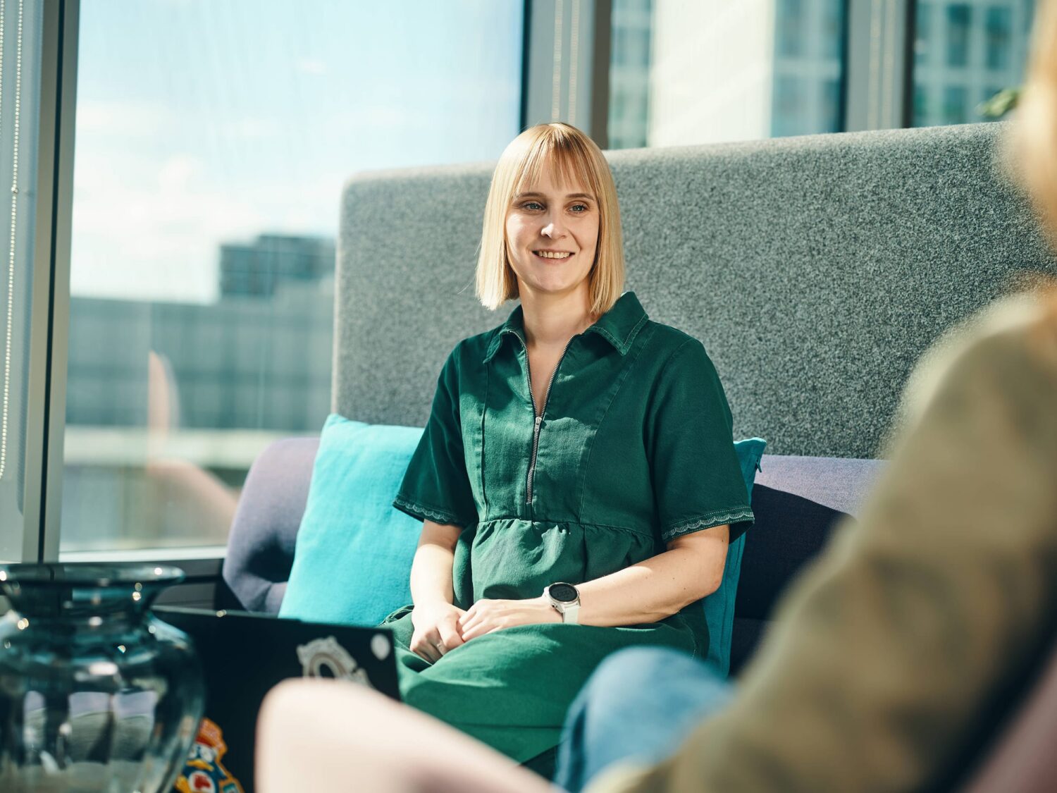 Smiling blonde woman in a green outfit sits on a sofa in a bright, modern office lounge with a laptop nearby.