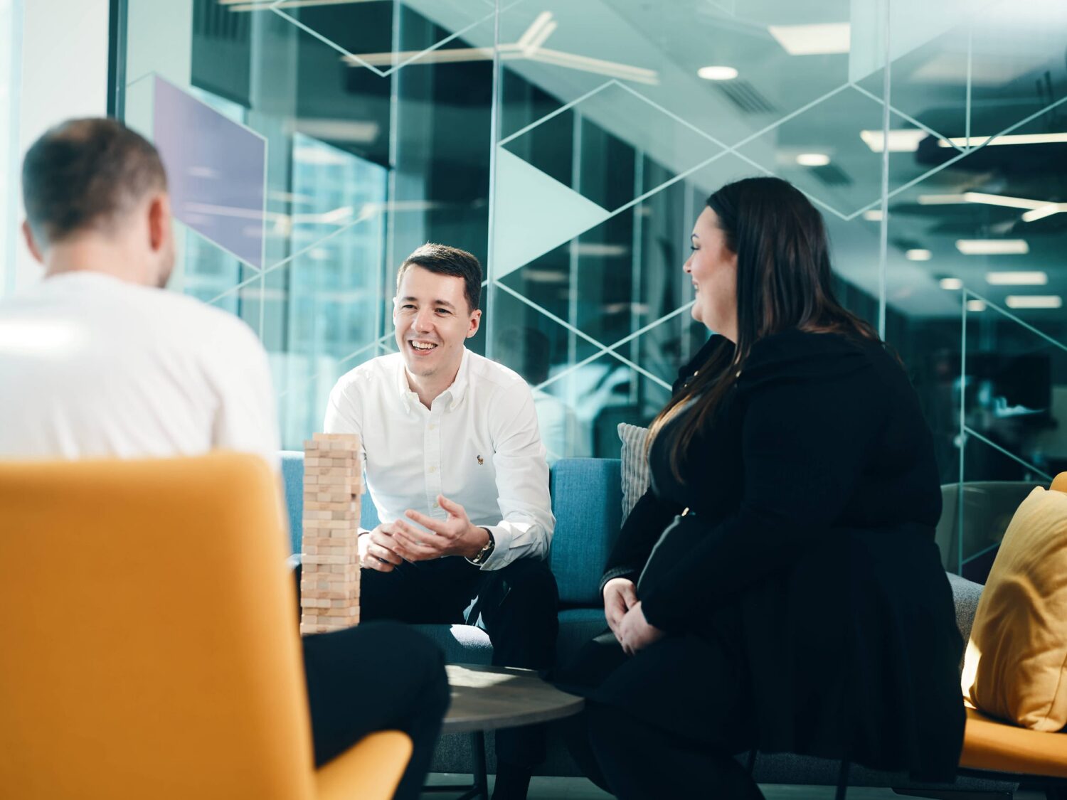 A group of coworkers in a modern glass-walled office chat around a small table with a wooden block tower.
