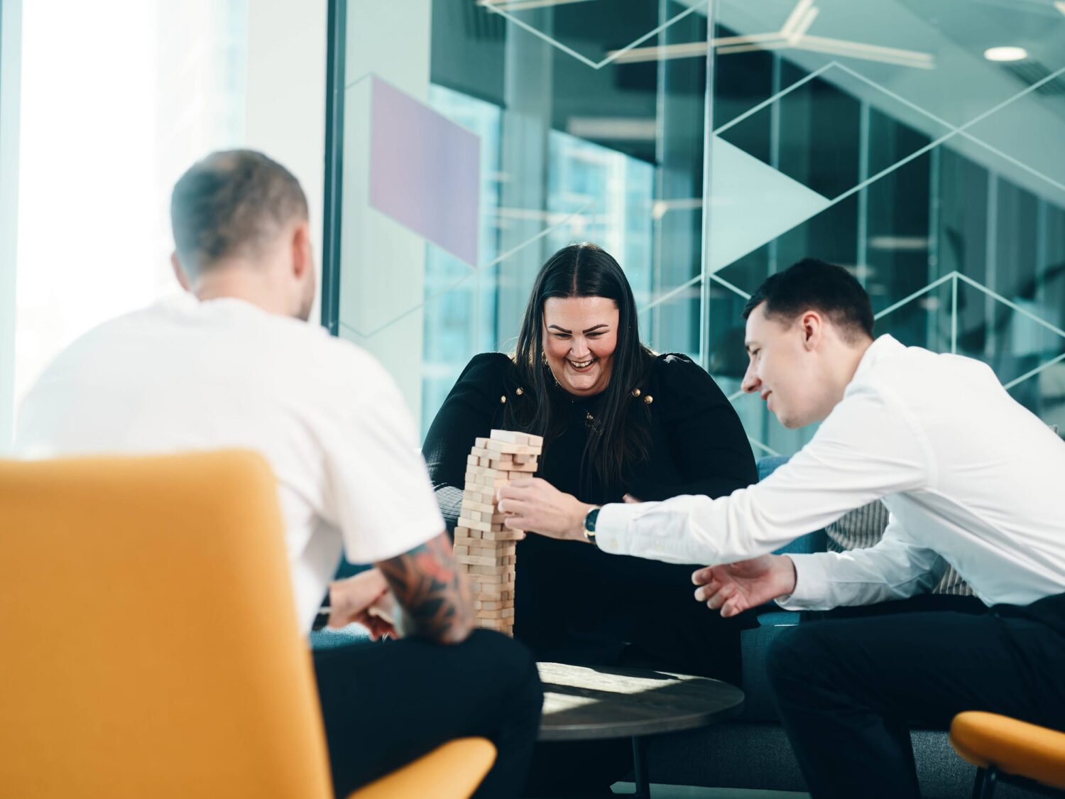 Three colleagues in a modern glass-walled office gather around a table, smiling at a smartphone.