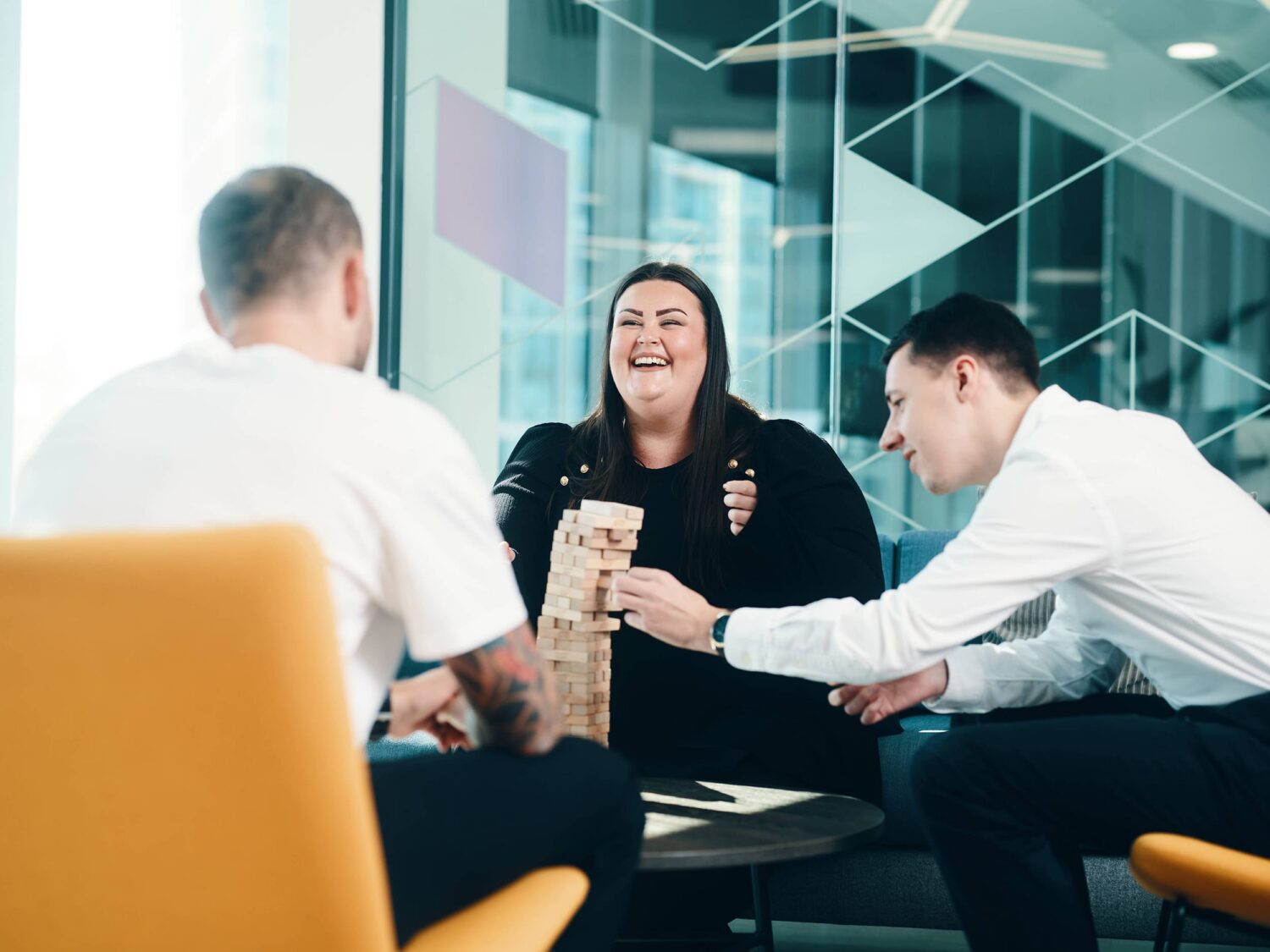 Three coworkers in a modern glass-walled office play Jenga, with a smiling woman at the center.