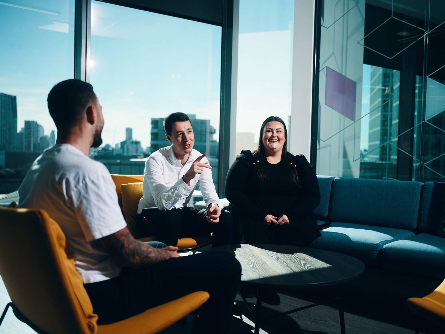Three people sit and chat in a modern office lounge with a city skyline visible through large windows.