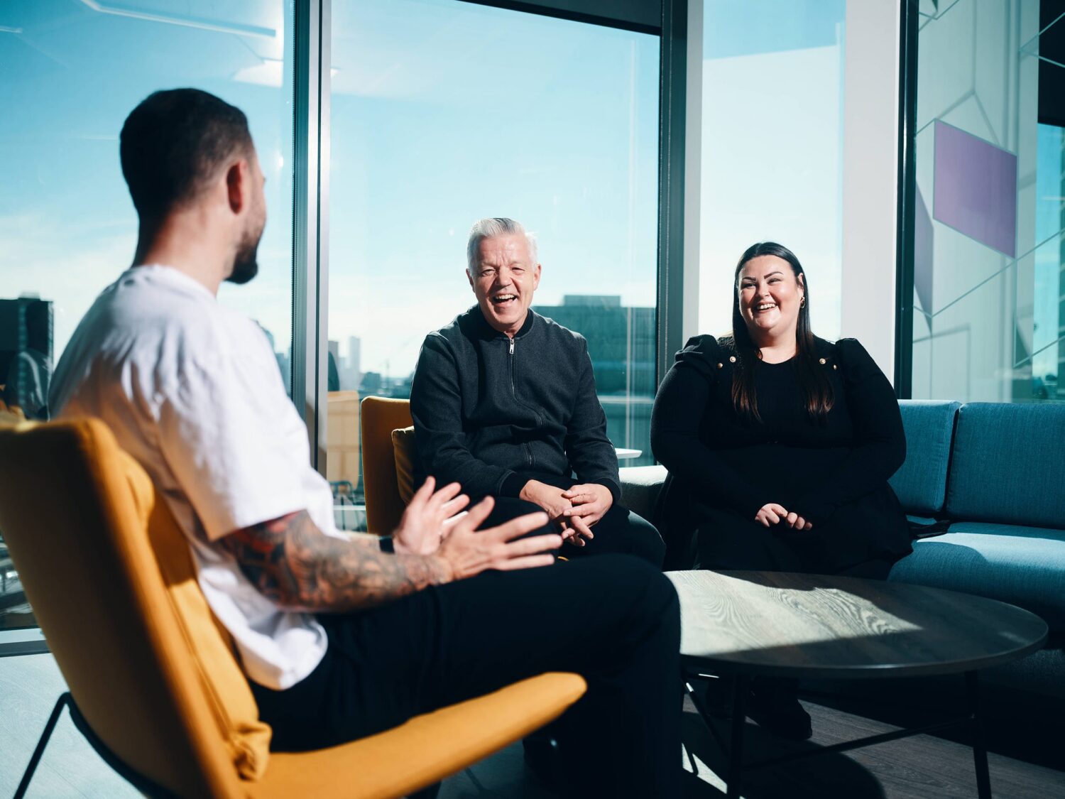 Three people sit in a modern glass-walled office lounge, engaged in conversation.
