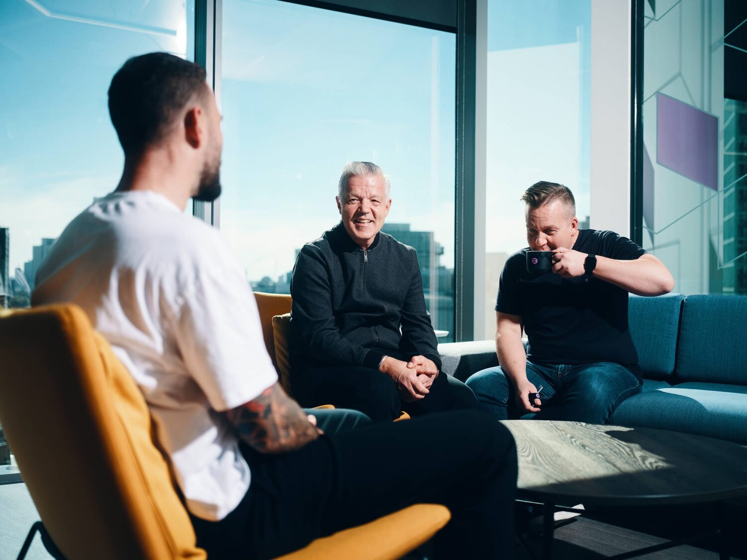 Three men sit in a bright glass-walled office lounge, engaged in conversation.