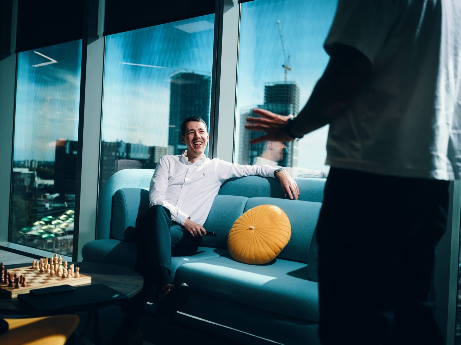 A man sits on a blue sofa with a yellow cushion in a modern glass-walled living room, while another person stands nearby and a chessboard rests on a table with a city view outside.