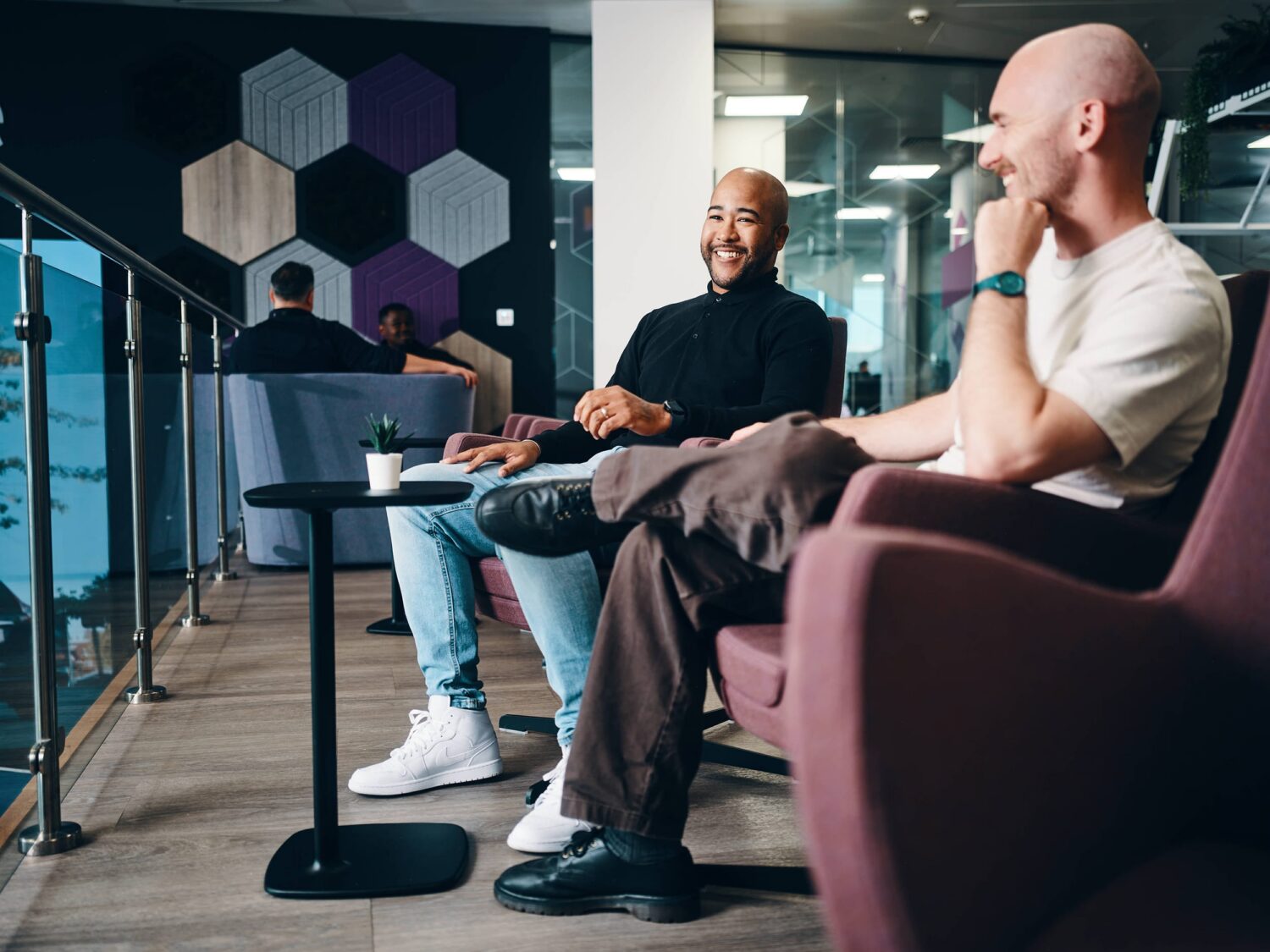 Two men sit and chat in a modern office lounge with glass railings and hexagonal wall panels.