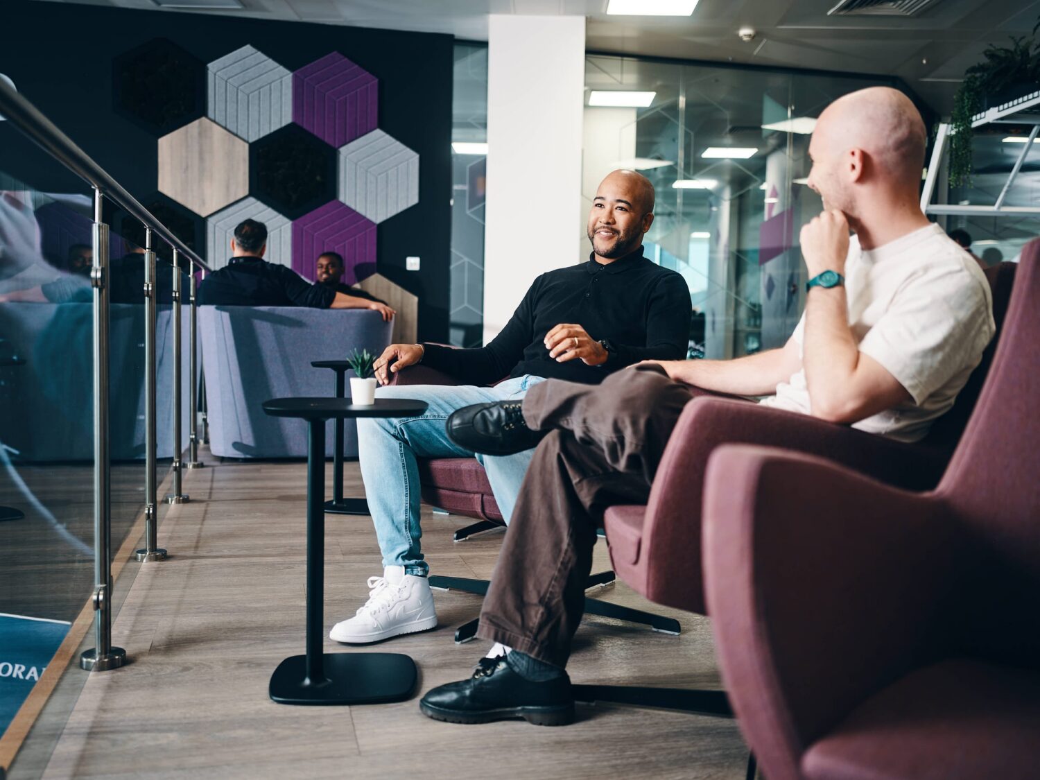 Two men are seated in a modern coworking space, talking beside a small round table among purple chairs and hexagonal wall panels.