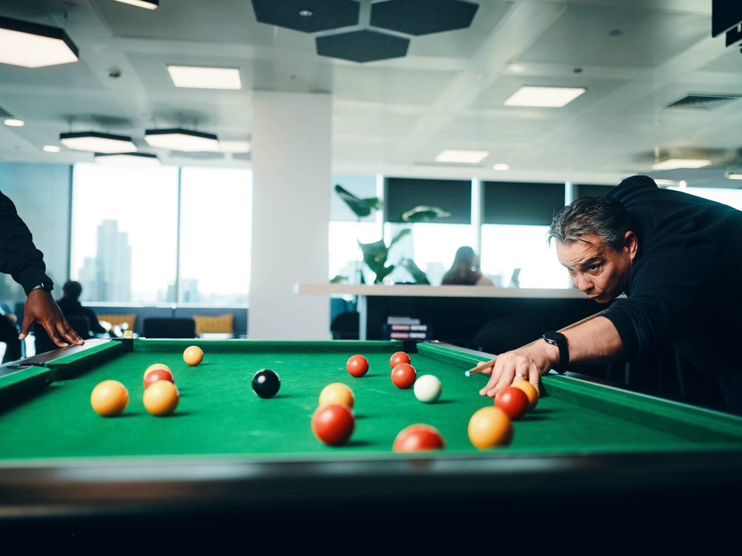 A man leans over a pool table, lining up a shot while colored balls rest on the green felt in a bright, modern room.