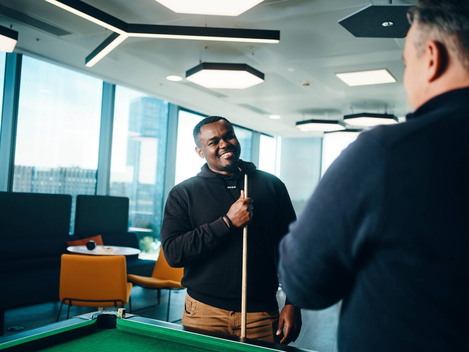 Two men stand near a pool table in a bright, modern lounge, with one man speaking into a microphone.