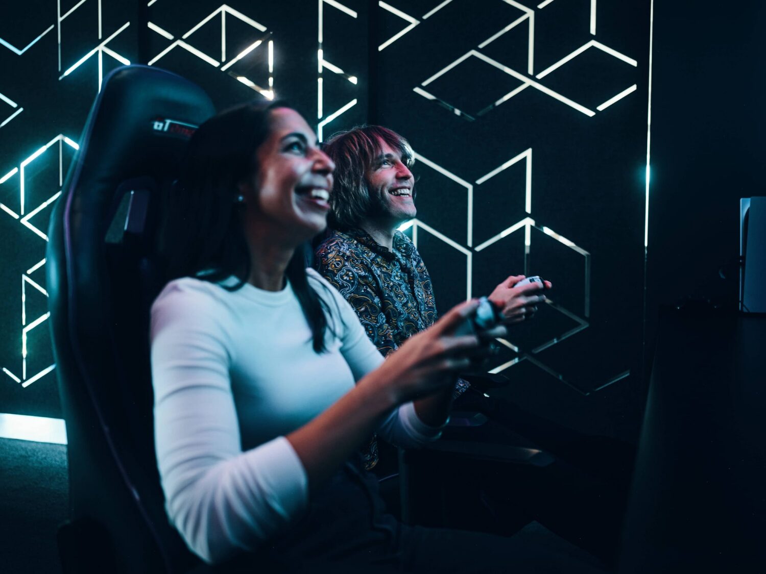 Two people sit in neon-lit arcade chairs, smiling as they play a virtual reality game.