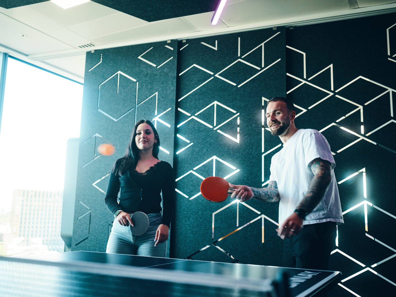 Two people playing table tennis indoors, a woman on the left and a man on the right, with a geometric-patterned wall in the background.