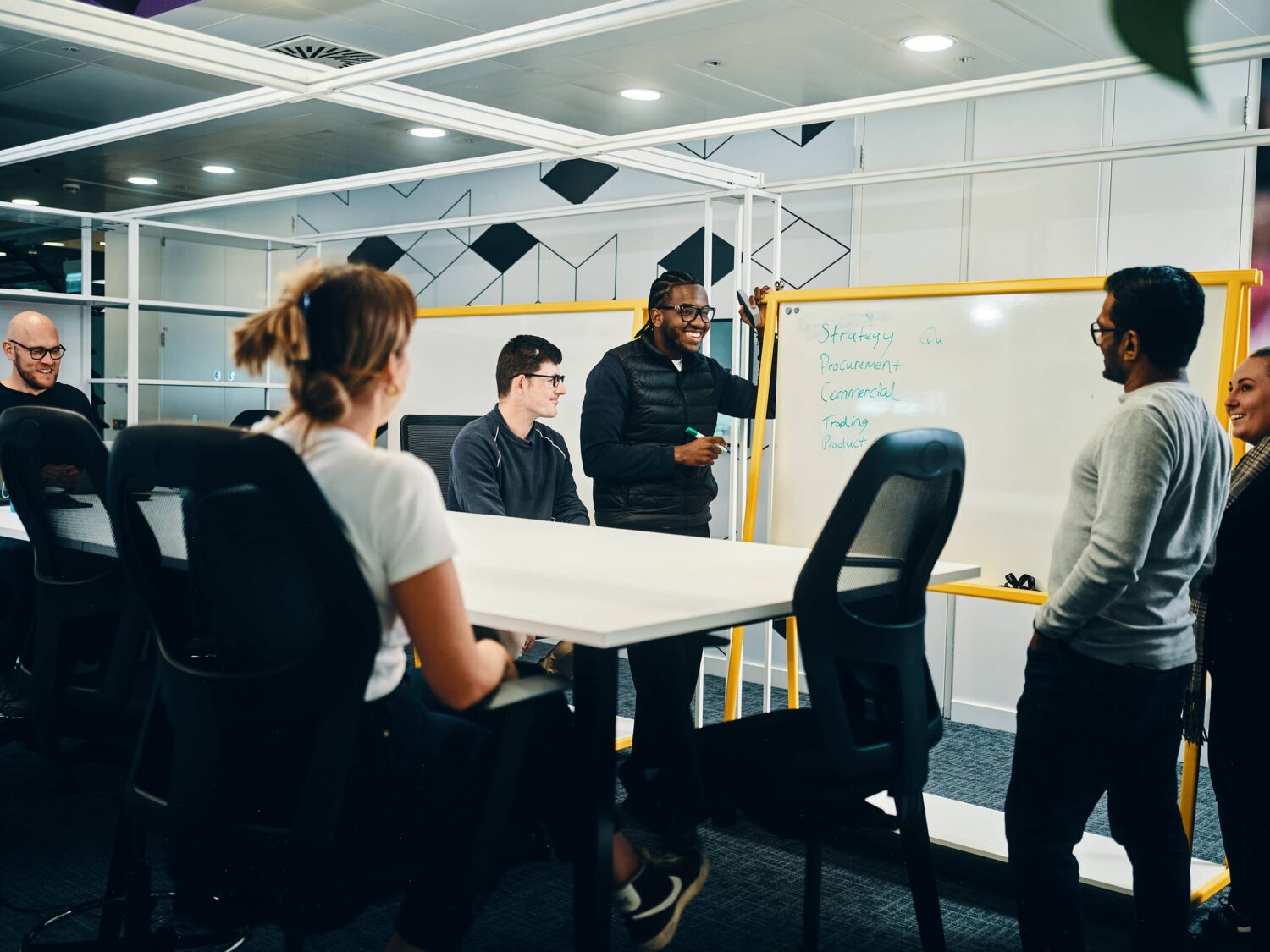 A group of people gather around a conference table in a modern office space with a yellow-framed whiteboard in the background.