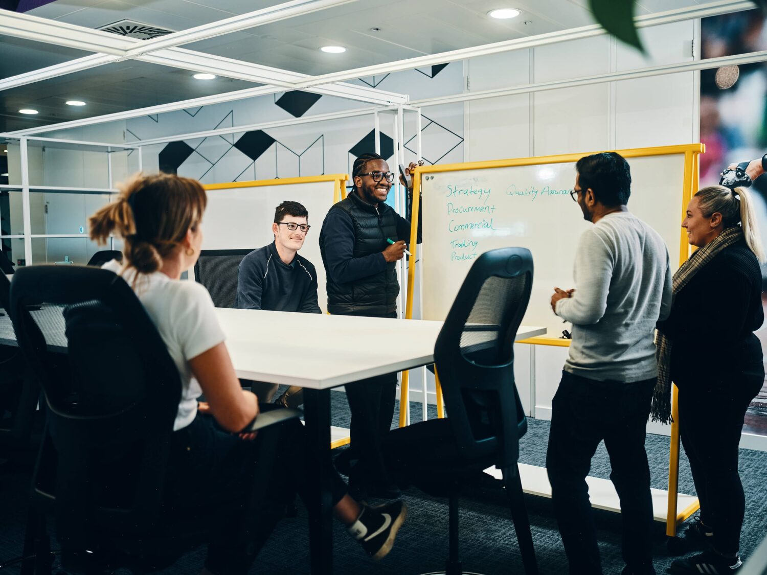 A group of people in a modern meeting room gathered around a conference table near a yellow-framed whiteboard.