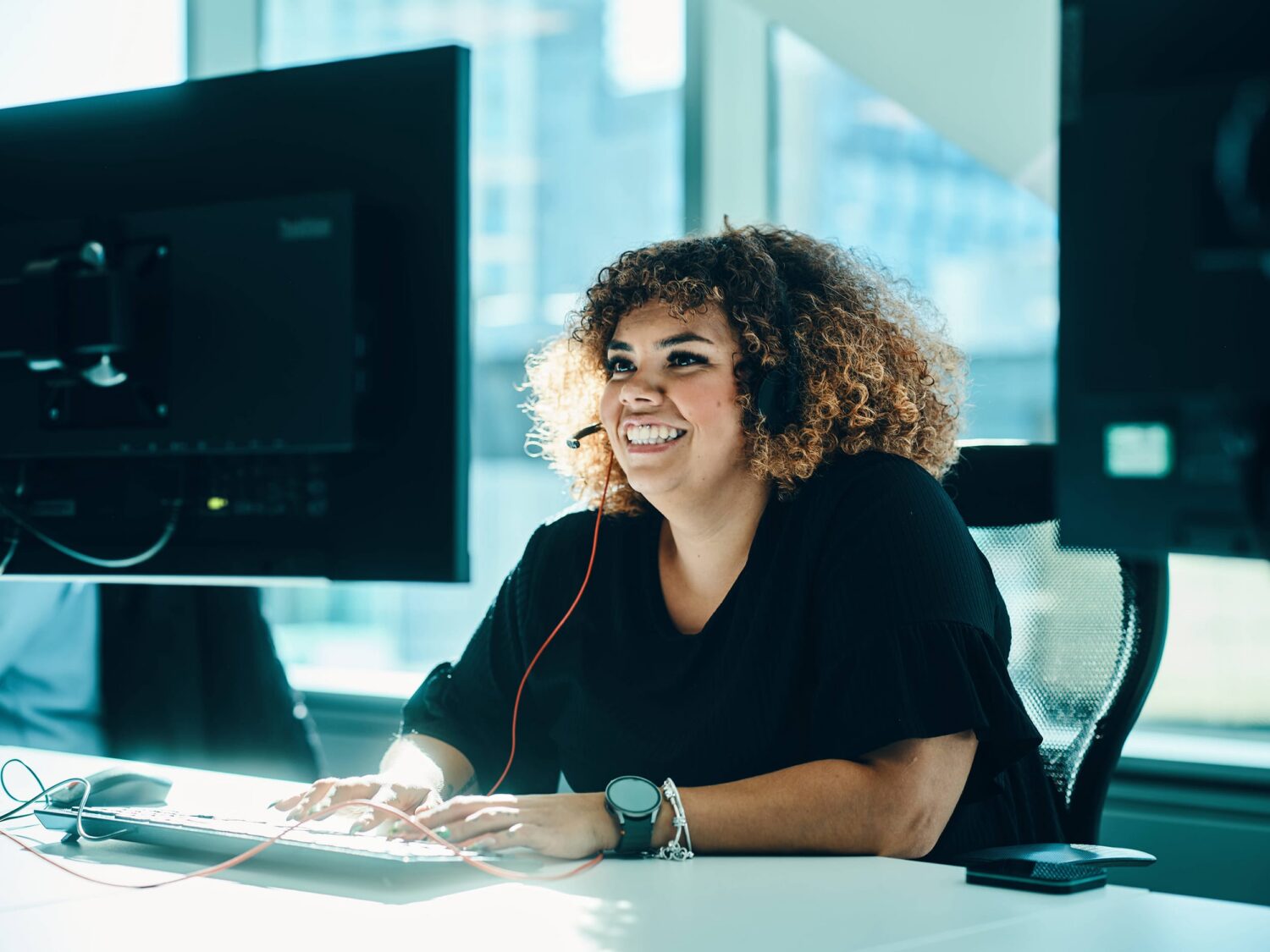 A woman with curly hair sits at a desk in an office, smiling while working at a computer.