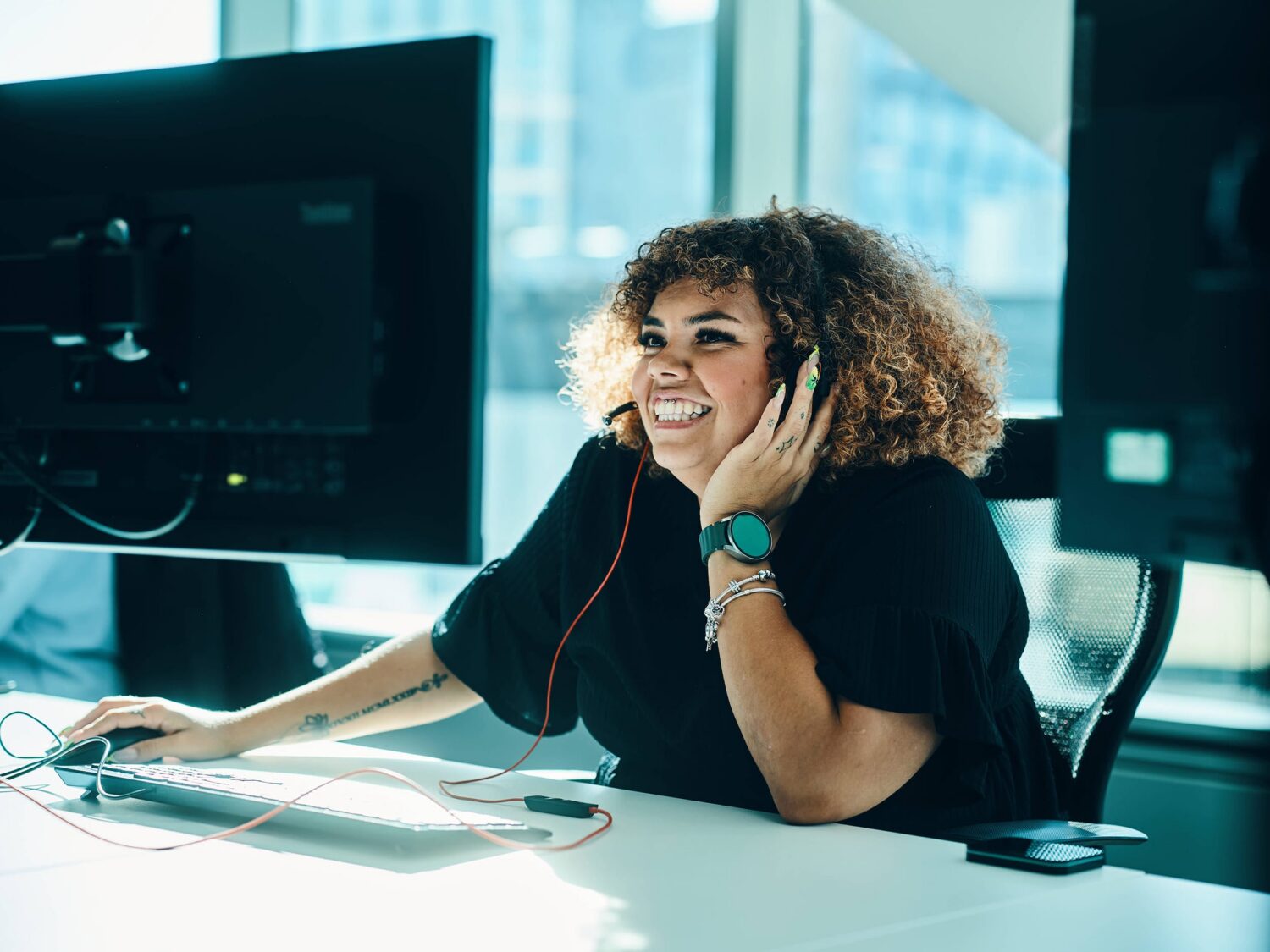 A smiling woman with curly hair wearing headphones sits at a desk in an office, using a computer.