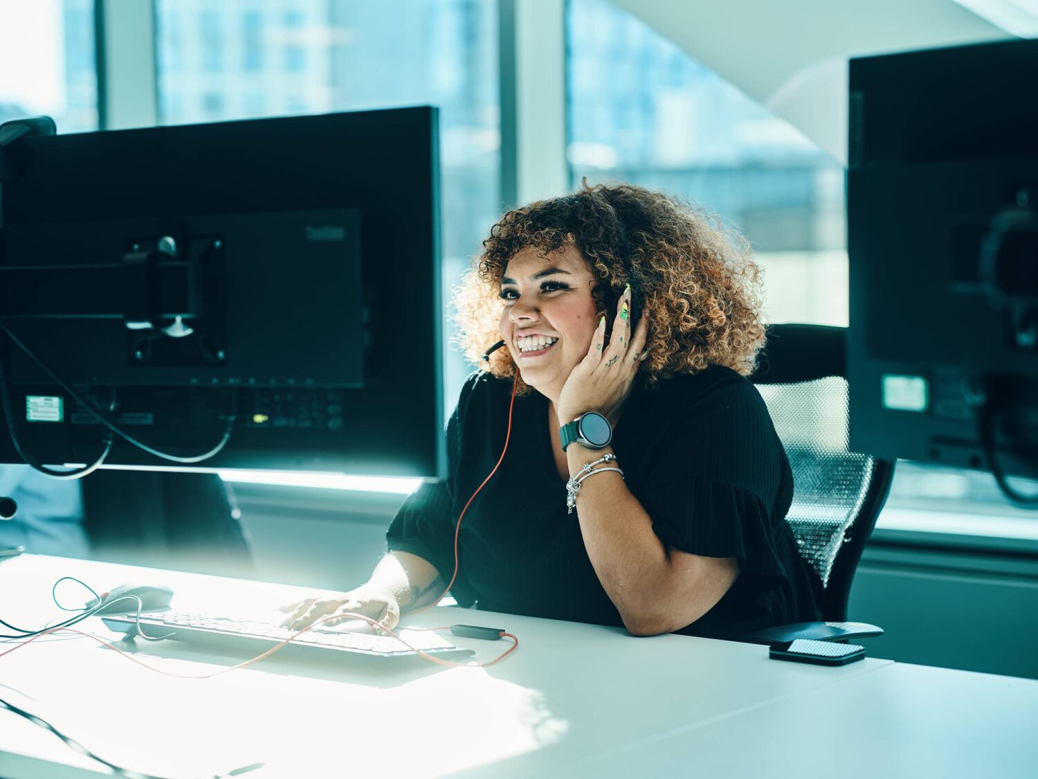 Smiling woman with curly hair wearing headphones sits at a bright desk with two computer monitors in an office.